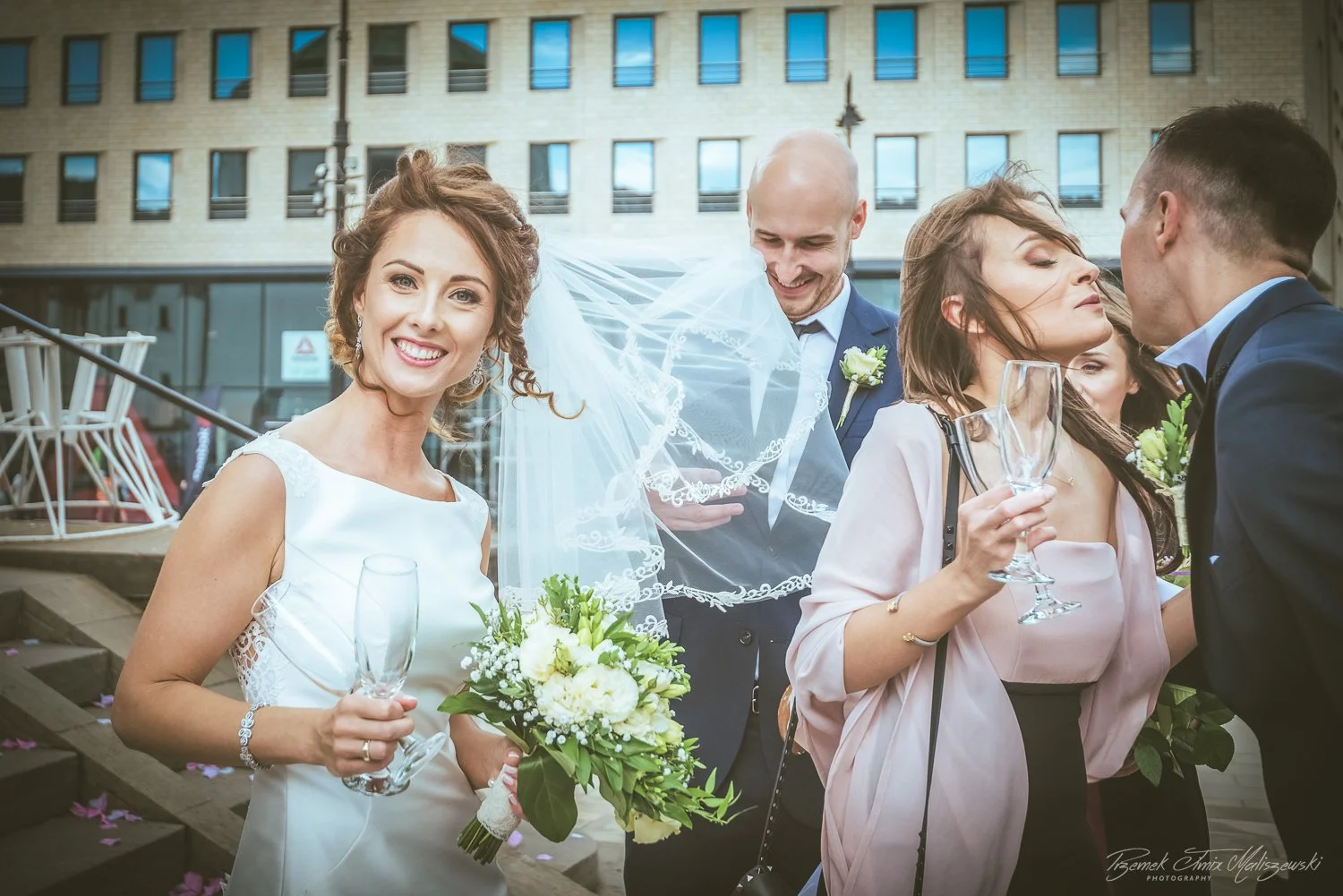 A bride in a white dress holding a bouquet and a champagne glass, smiling at the camera during her wedding celebration, with guests behind her, some holding champagne glasses and engaging in conversation, outdoor urban background.
