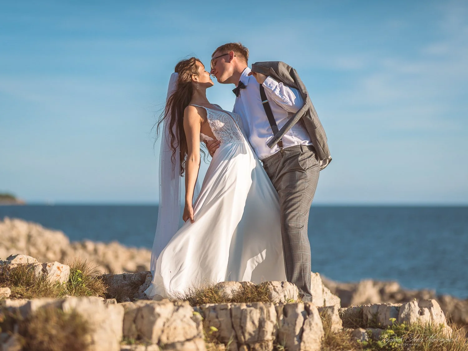 A bride and groom share a kiss on a rocky coastal area, with the ocean and blue sky in the background, during their wedding photoshoot.