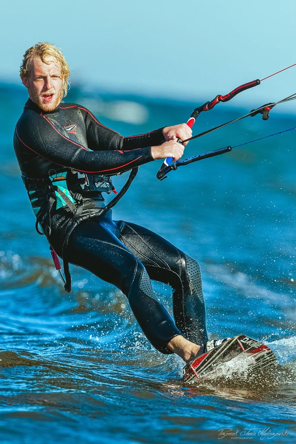 Man riding a wakeboard on the water, wearing a black wetsuit and holding onto a tethered handle, water splashing around.