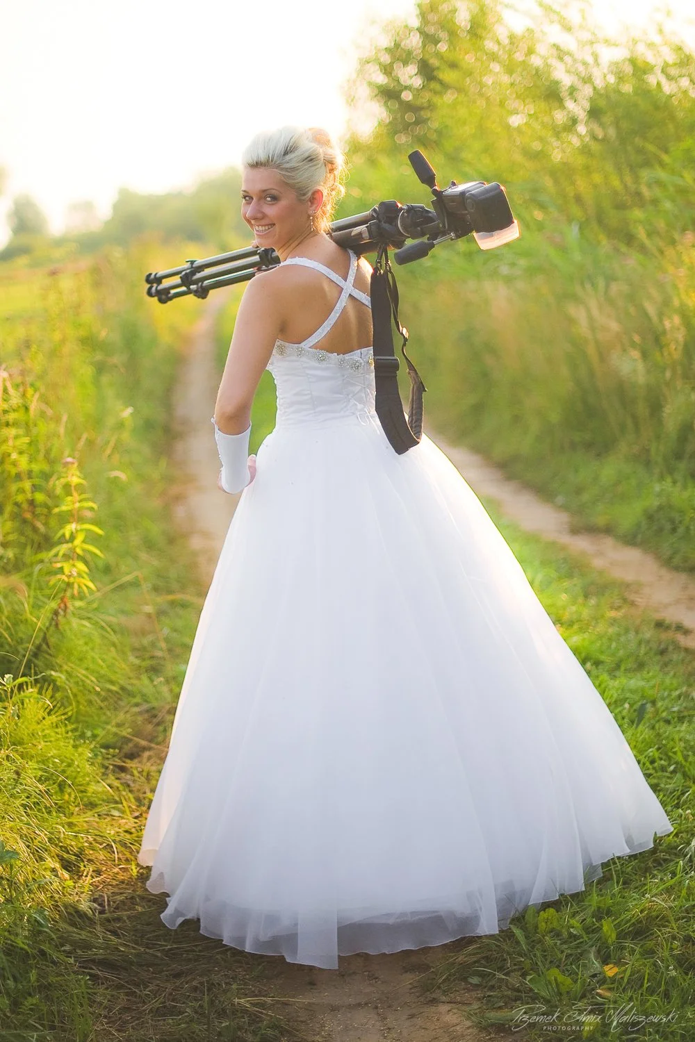 A smiling woman in a white wedding dress and gloves carrying a camera tripod on her shoulder, standing on a dirt path surrounded by green vegetation during sunset.