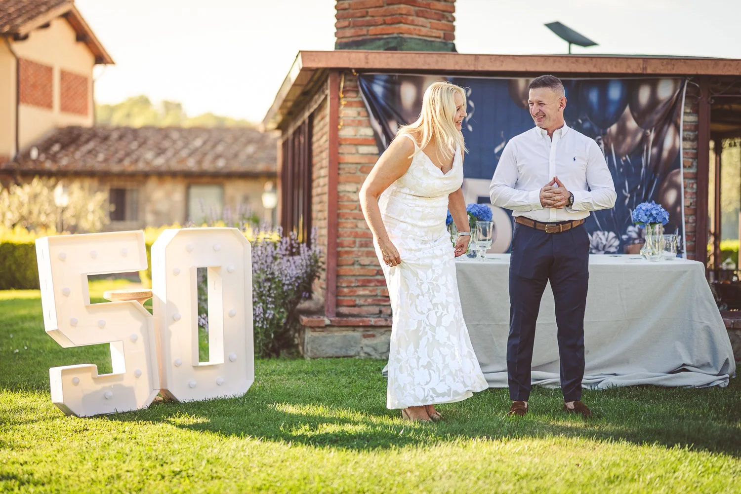 A woman and a man are standing outdoors in front of a decorated table. The woman, in a white dress, is smiling and looking at the man, who is dressed in a white shirt and dark pants. There is a large illuminated "50" sign on the grass, indicating a 50th celebration, likely a birthday or anniversary.