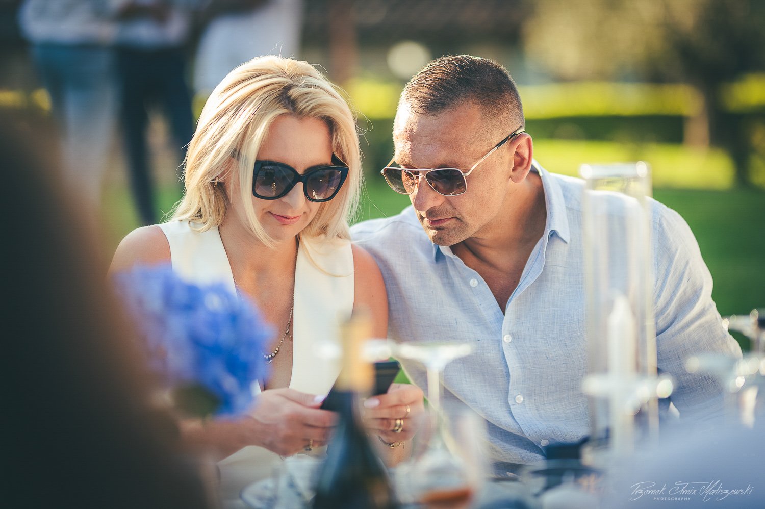 A man and woman wearing sunglasses looking at a phone while sitting outdoors at a table with drinks.