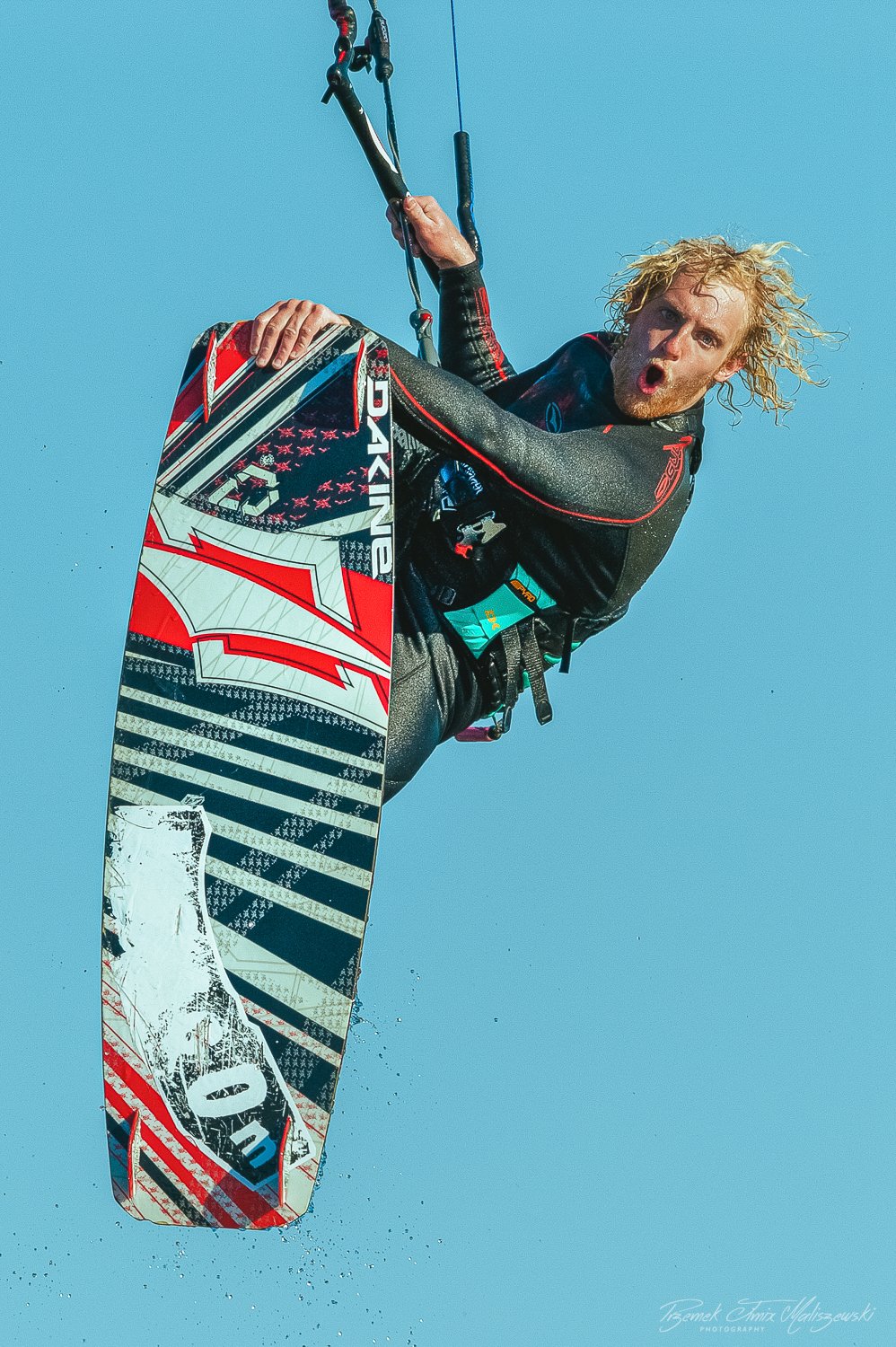 Kiteboarder flying in the air with a colorful kiteboard and blue sky background.