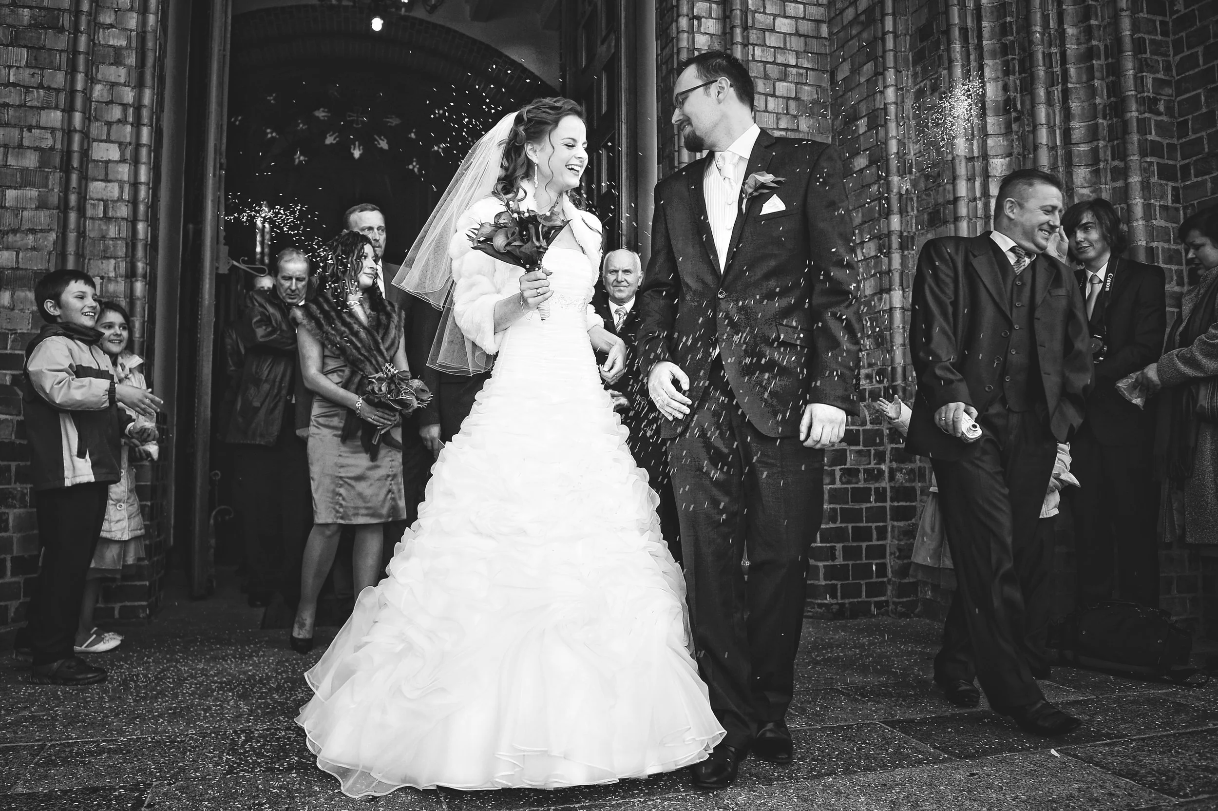 Black and white photo of a wedding celebration with a bride and groom smiling at each other, surrounded by guests outside a brick building, with confetti falling.