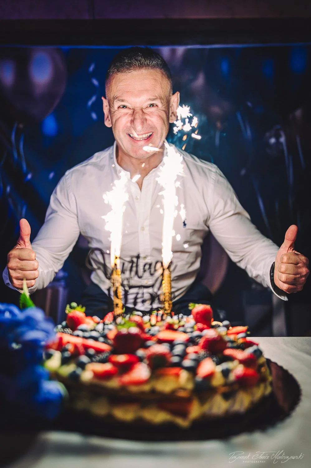 A man celebrating birthday with a large cake topped with strawberries and blueberries, with sparklers on the cake, giving a thumbs-up and smiling.