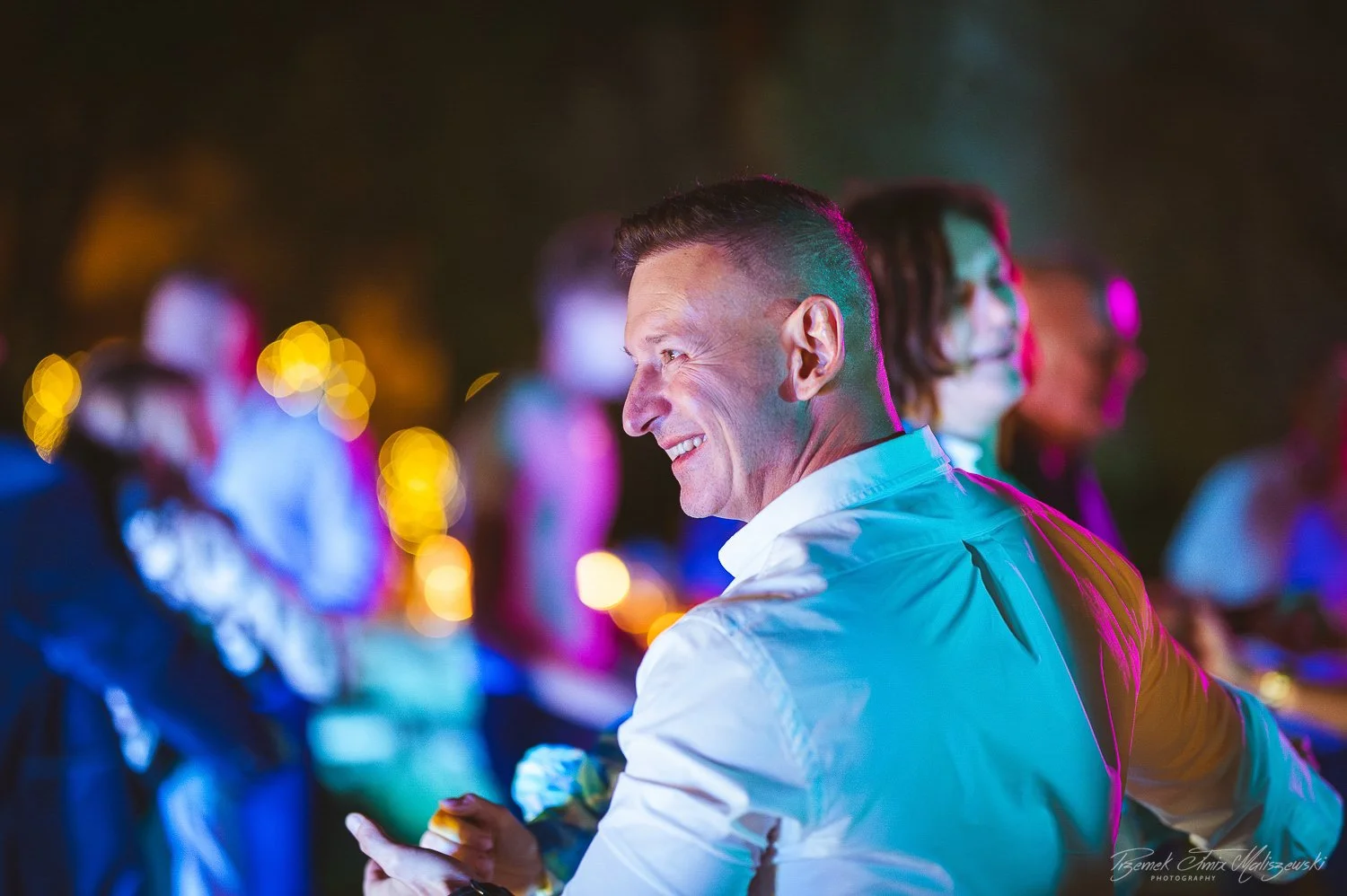 A man in a white shirt smiling and dancing at a lively party or celebration, with colorful blurred lights and other people in the background.