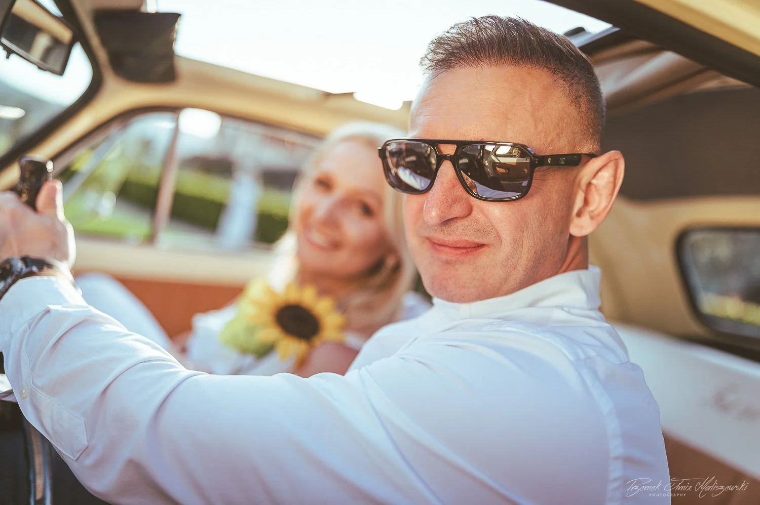 A man wearing sunglasses and a white shirt sitting in the driver's seat of a vintage car, with a woman smiling in the background holding a sunflower.