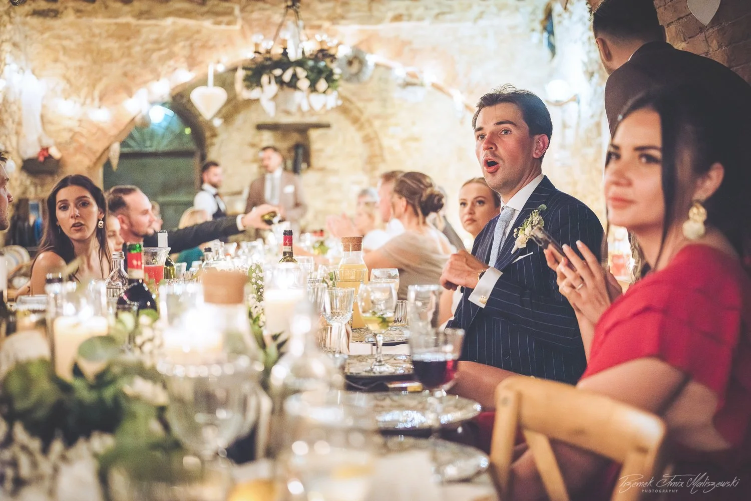 People attending a wedding reception sitting at a decorated long table with drinks, florals, and candles in a rustic venue with exposed brick walls and romantic lighting.
