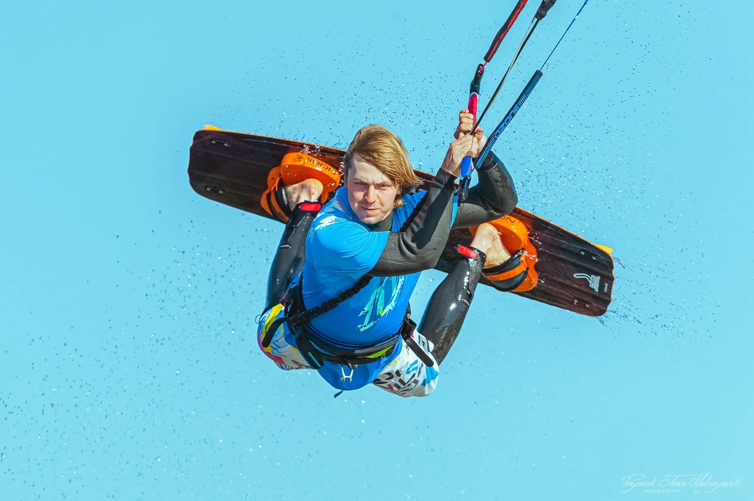 Kiteboarder in mid-air performing a jump over water with a blue sky background, holding onto a kite's control bar.