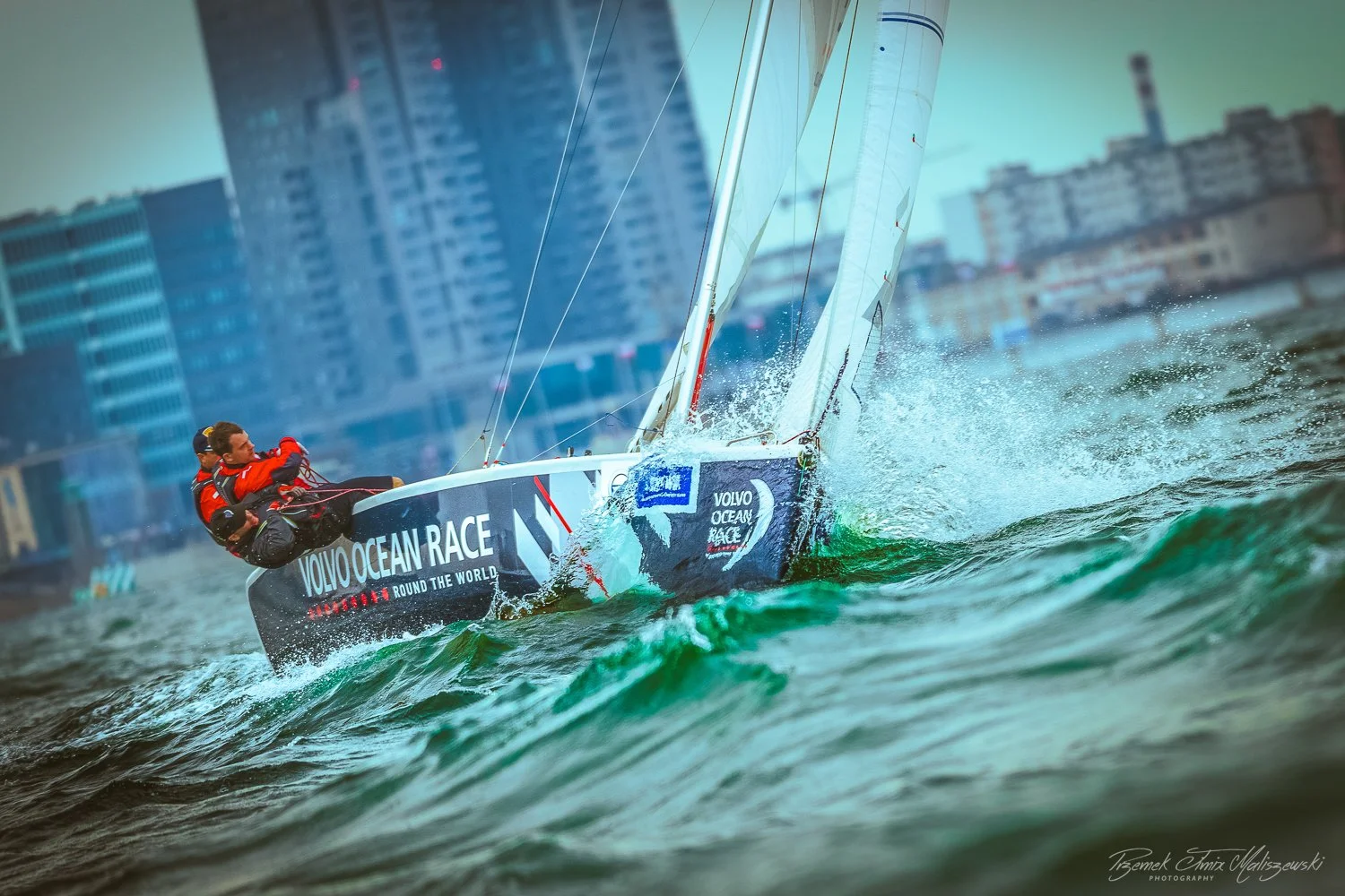 Sailing boat racing in the Volvo Ocean Race round the world, with crew members wearing red jackets and city buildings in the background