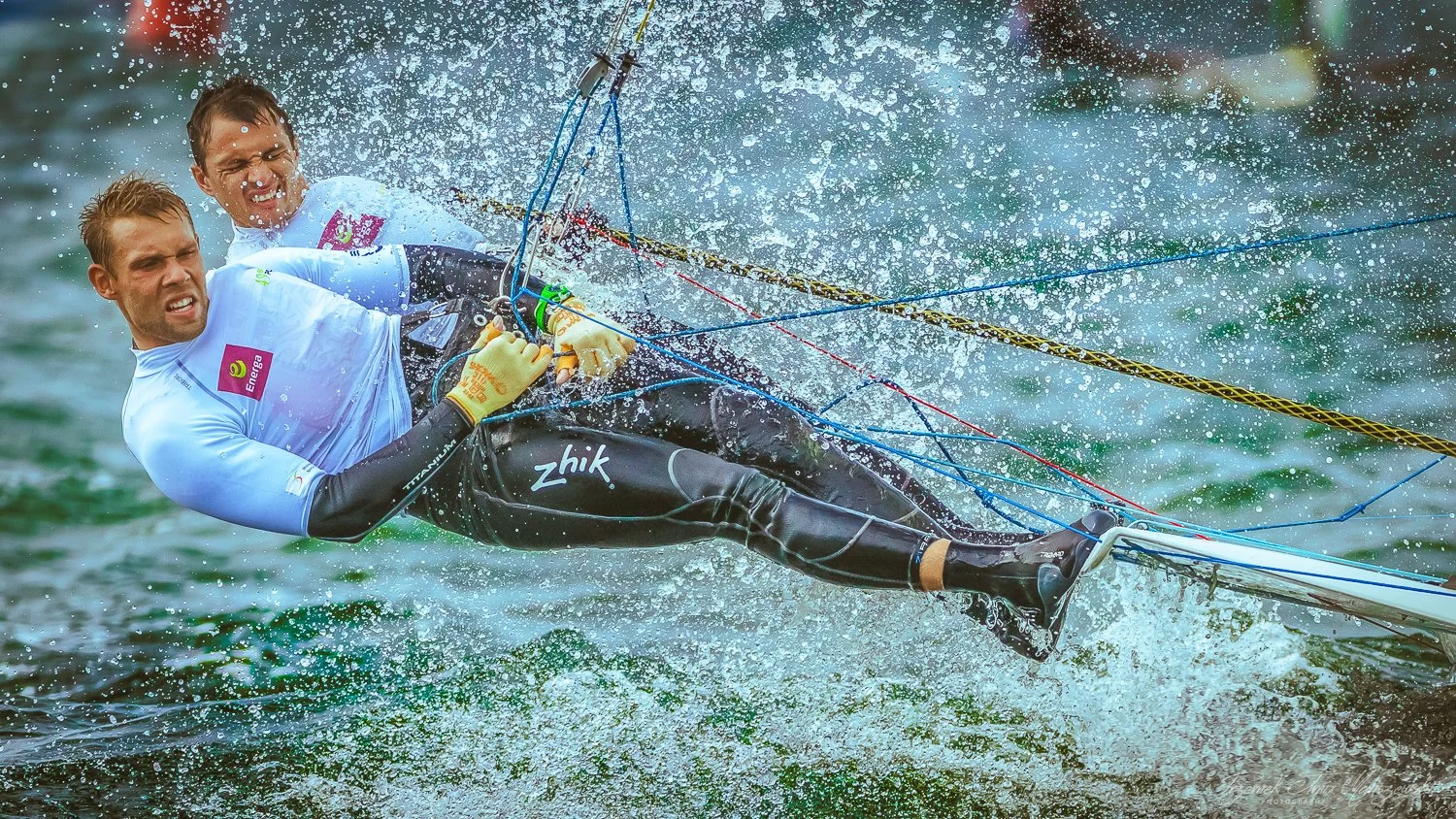 Two men in wetsuits and T-shirts are falling into water while holding onto a kiteboarding kite's control bar, creating a splash.