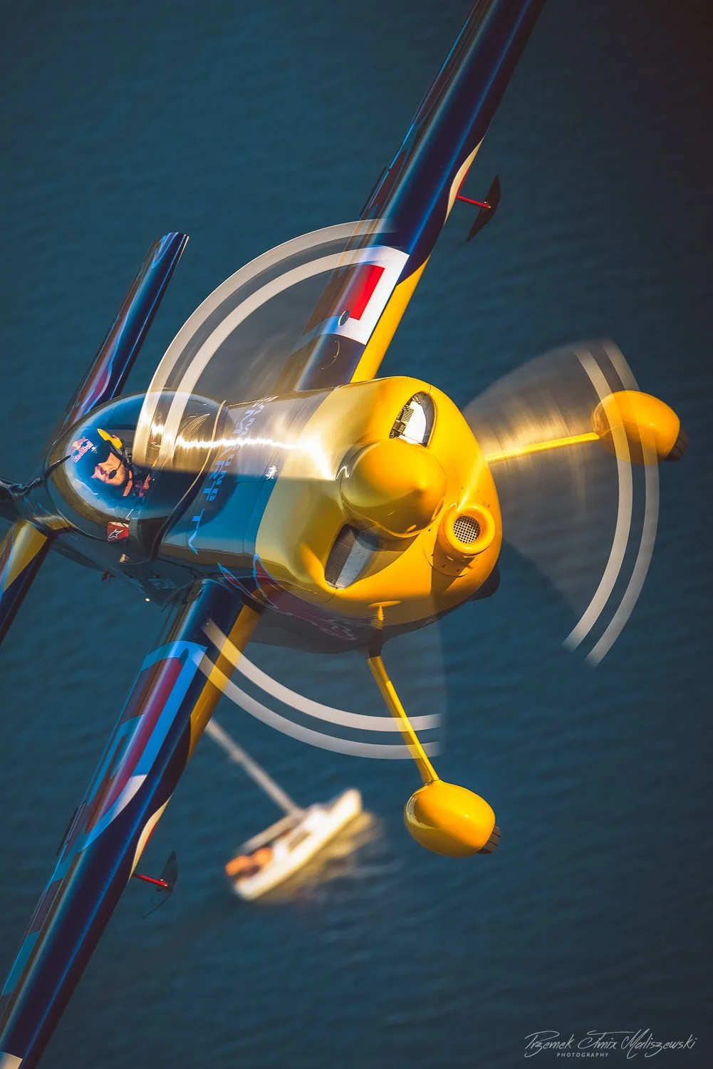 A yellow aerobatic aircraft flying low over water with a pilot inside and a boat visible in the background.