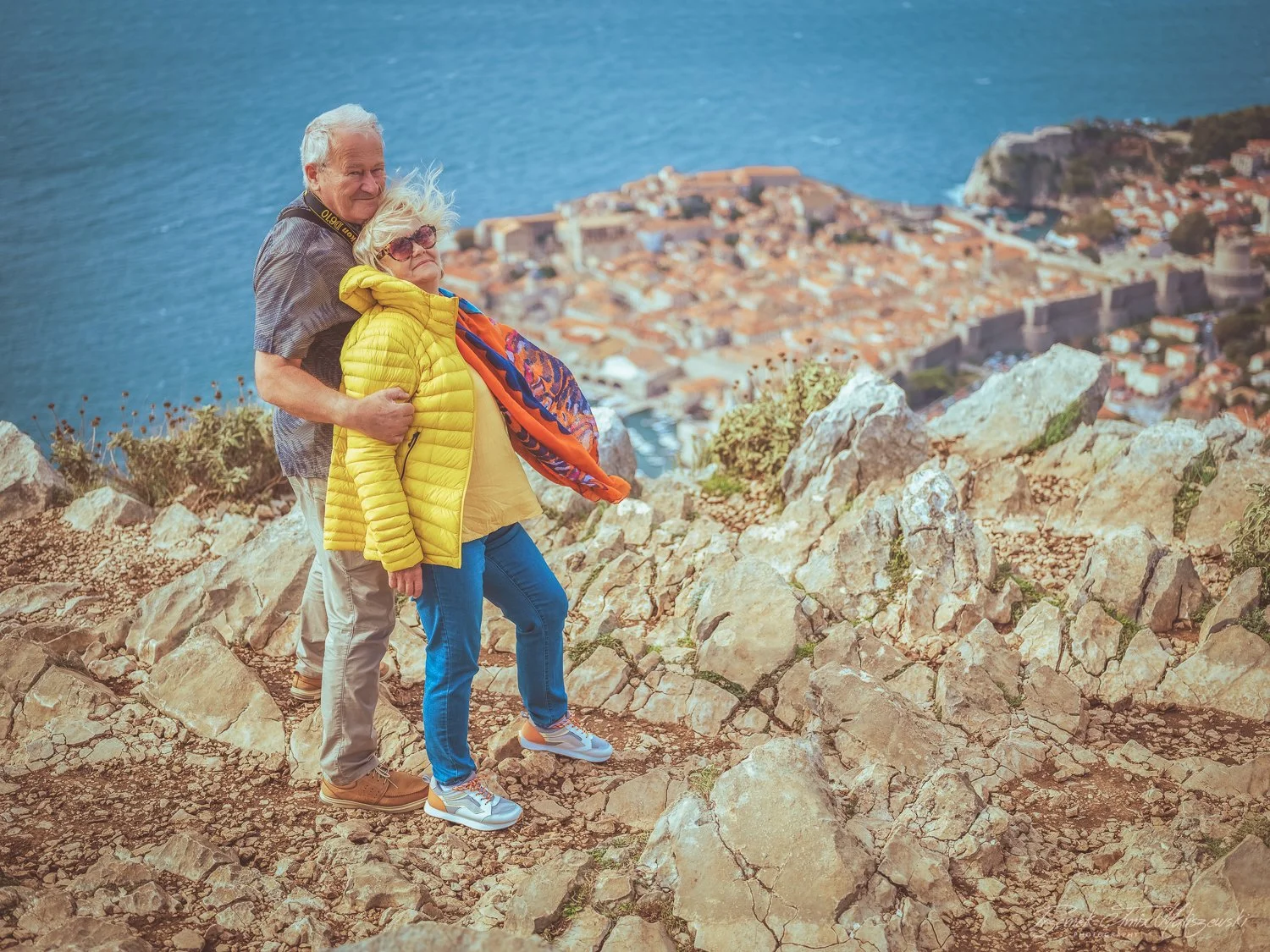 Two elderly people, a man and a woman, standing on rocky terrain overlooking a coastal town with orange rooftops and a large body of water in the background. The woman is wearing a bright yellow jacket, scarf, sunglasses, and blue jeans, while the ma