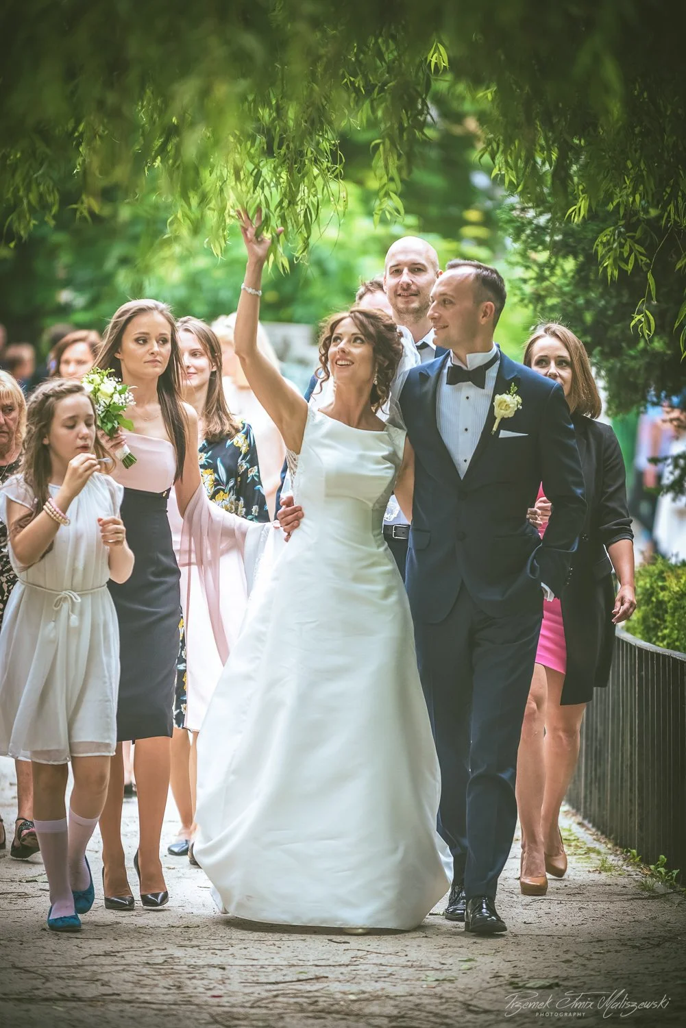 A bride and groom walking outdoors with friends and family, some holding flowers and dressed in formal attire.