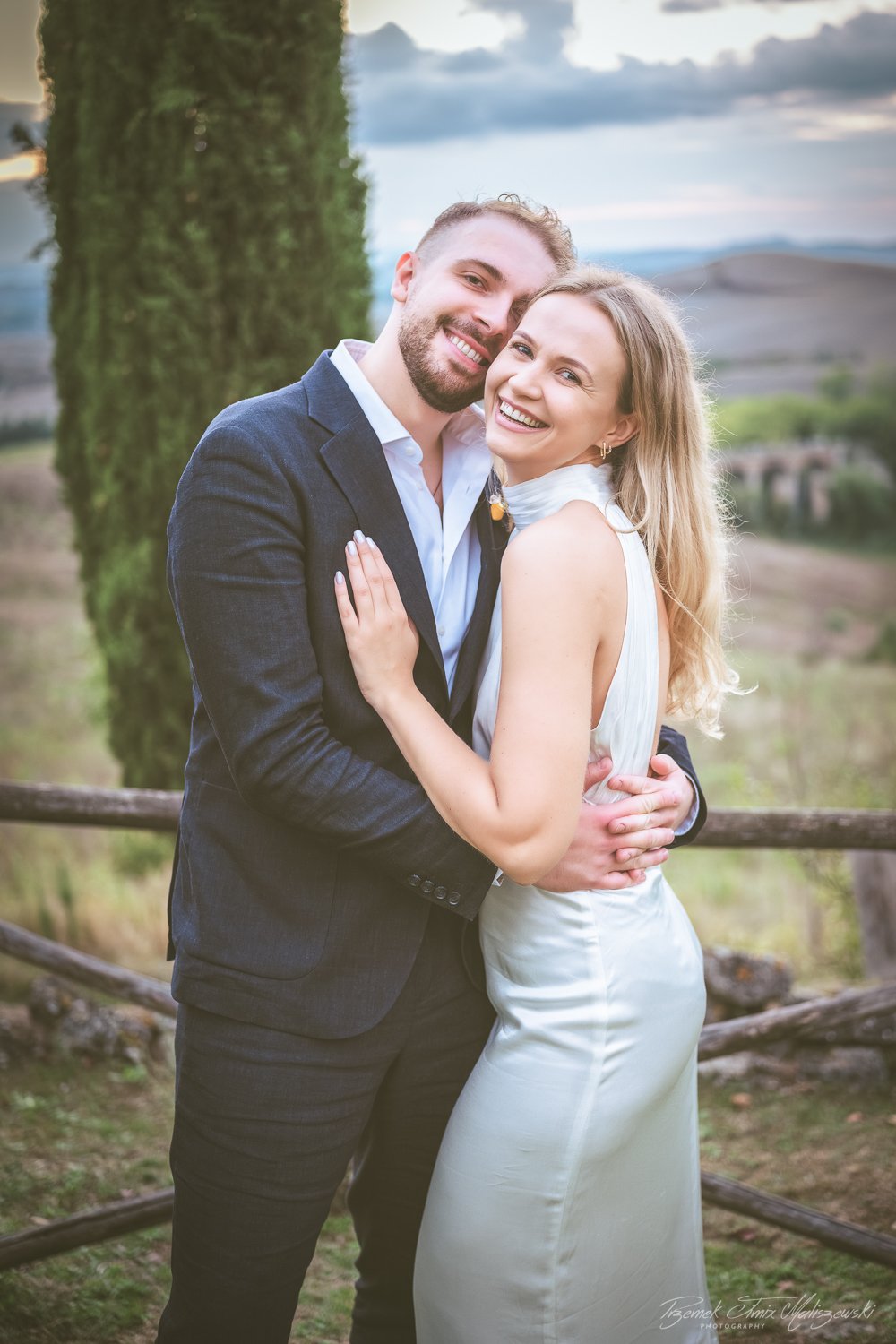 A happy couple embracing outdoors near a wooden fence with a rural landscape and cloudy sky in the background.