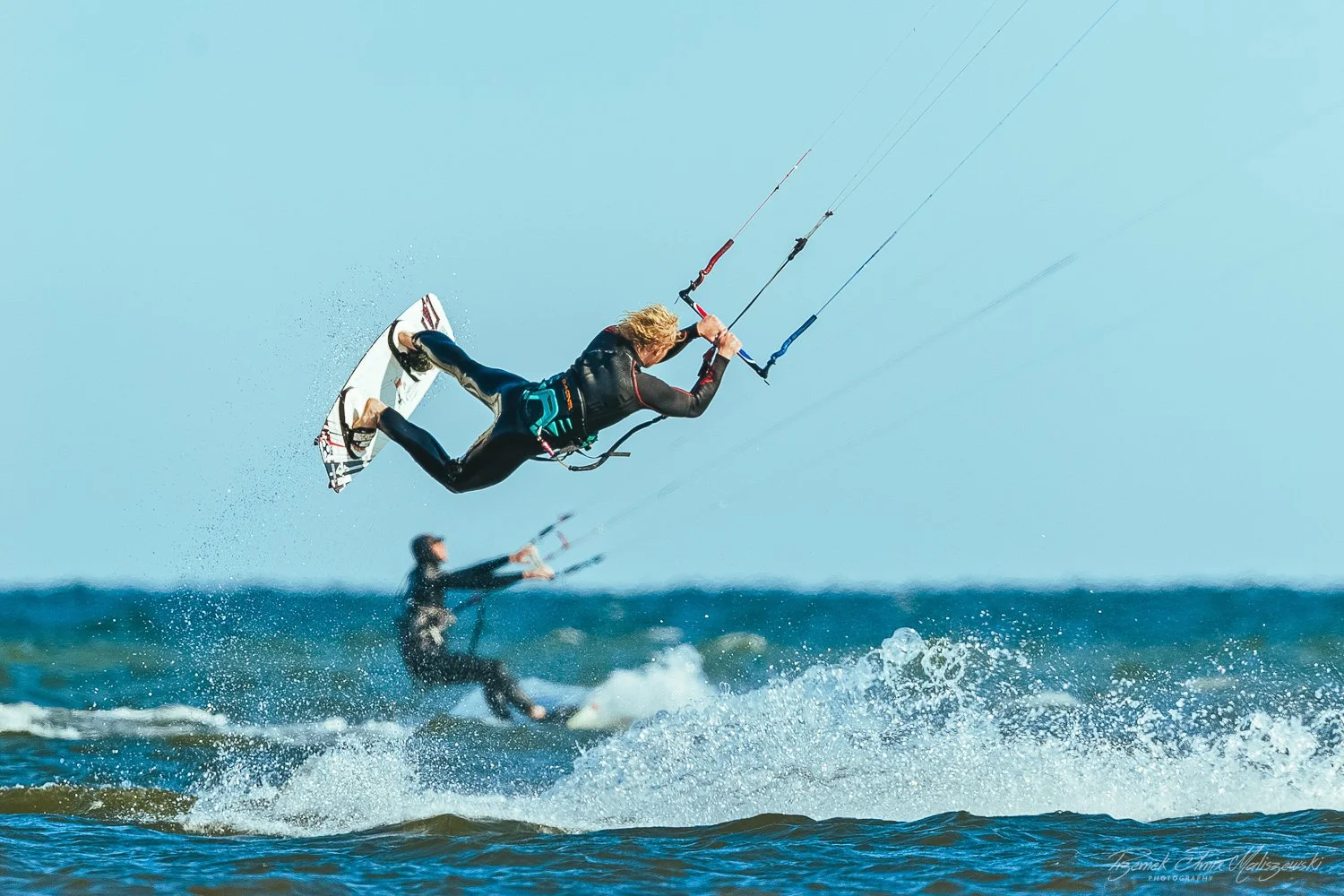 Two kiteboarders riding on the ocean with one jumping into the air. Both are wearing wetsuits.