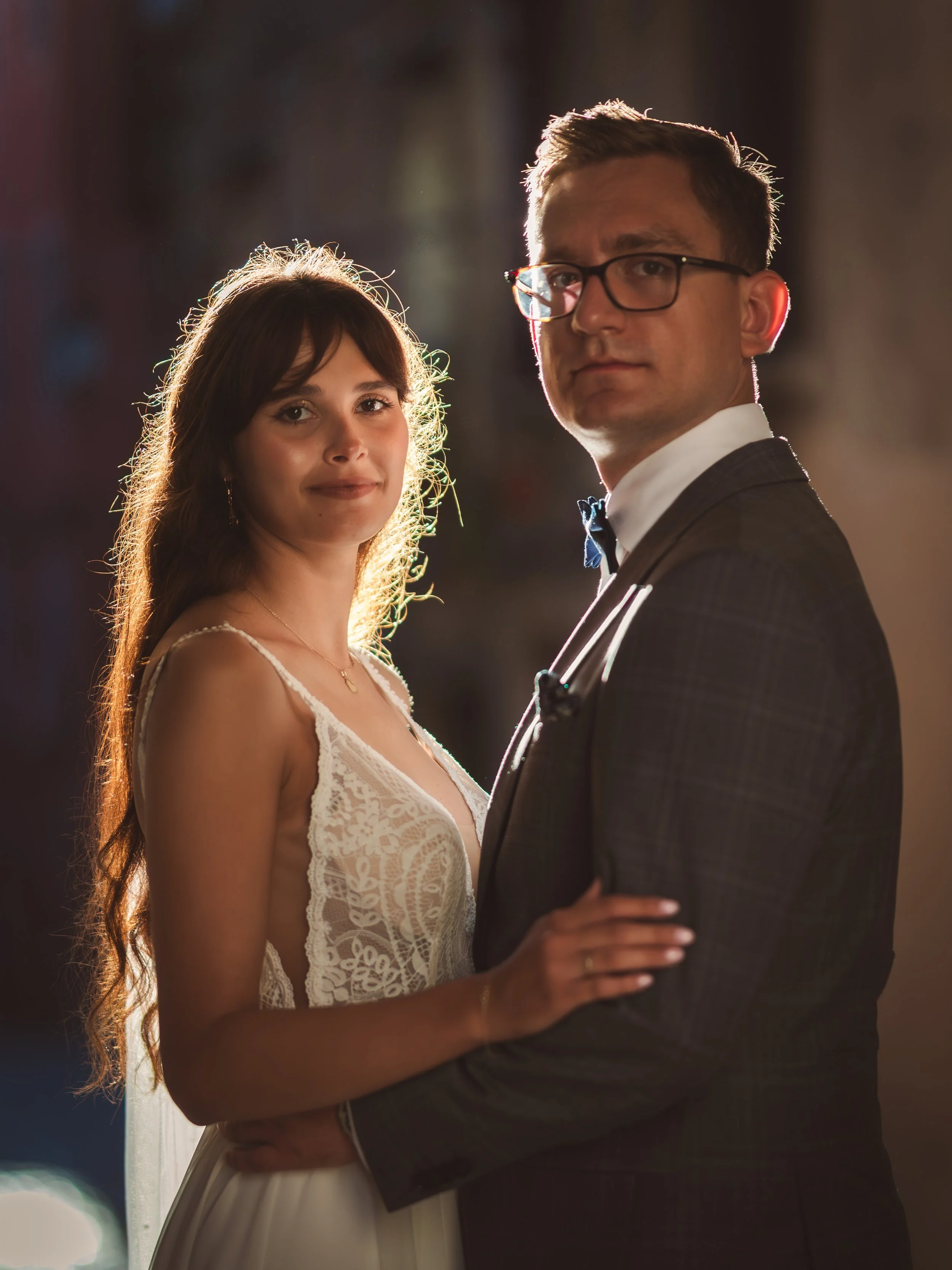 A young woman in a white lace dress and a man in a tuxedo, standing close together at a formal event or wedding, with warm lighting illuminating their faces.