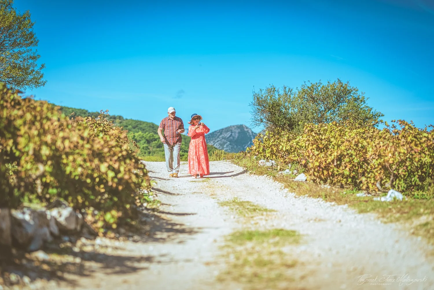 Two people, a man and a woman, walking along a dirt path surrounded by bushes and trees, with mountains in the background and a clear blue sky overhead.