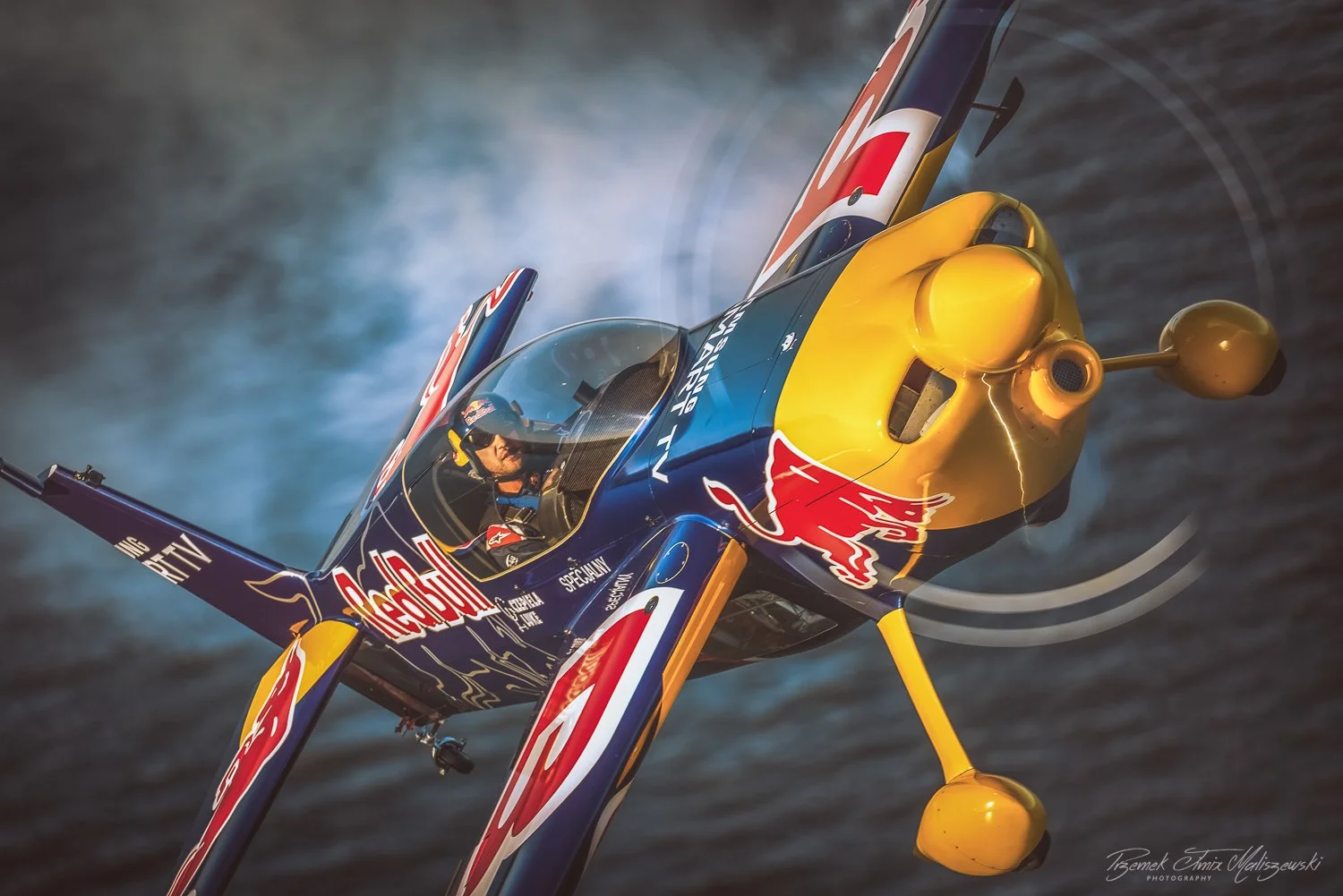 A Red Bull-sponsored aerobatic airplane flying low over water, with smoke trailing behind.