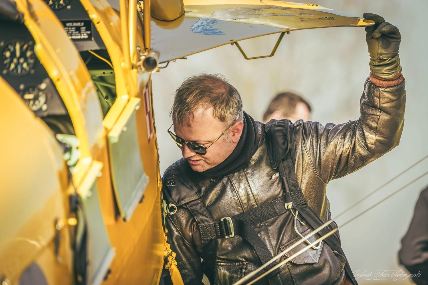 Man wearing sunglasses, leather jacket, and harness working on the yellow aircraft cockpit.