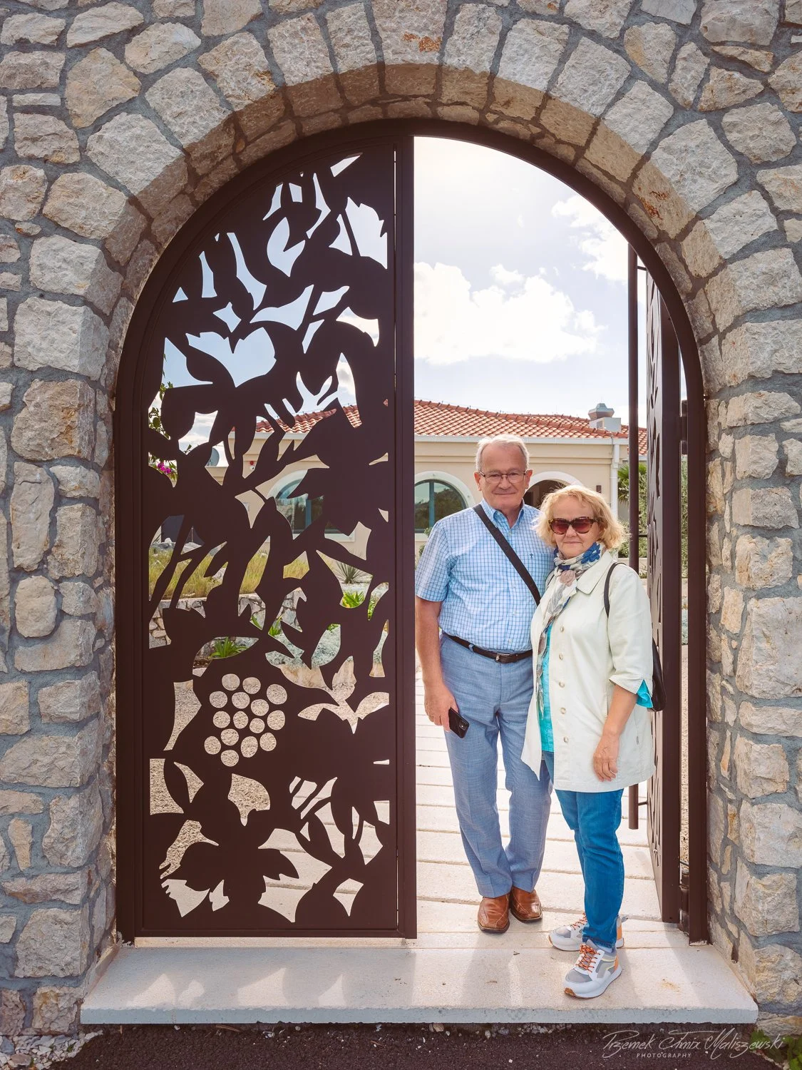 An elderly man and woman standing together in front of a decorative metal gate with a tree and grape cluster design, framed by a stone arch, outside a building with a red-tiled roof on a partly cloudy day.