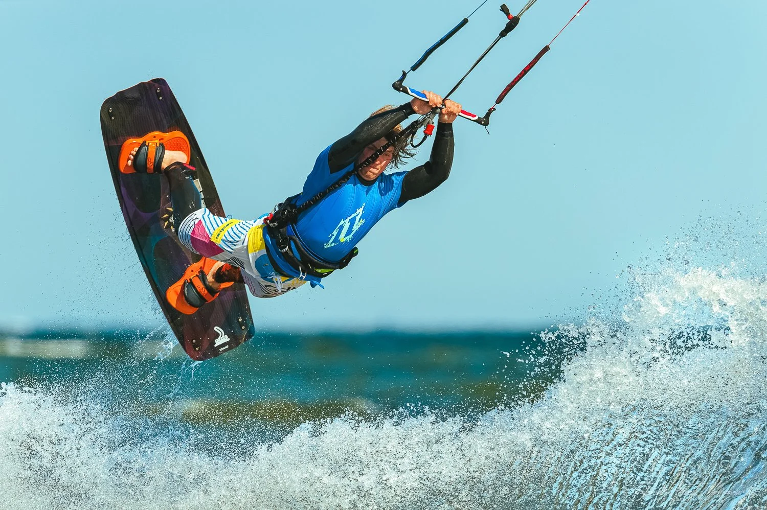 Kiteboarder performing an aerial trick above the water, wearing a blue shirt, multicolored shorts, and orange sandals, with a kite control bar in hand during the daytime.