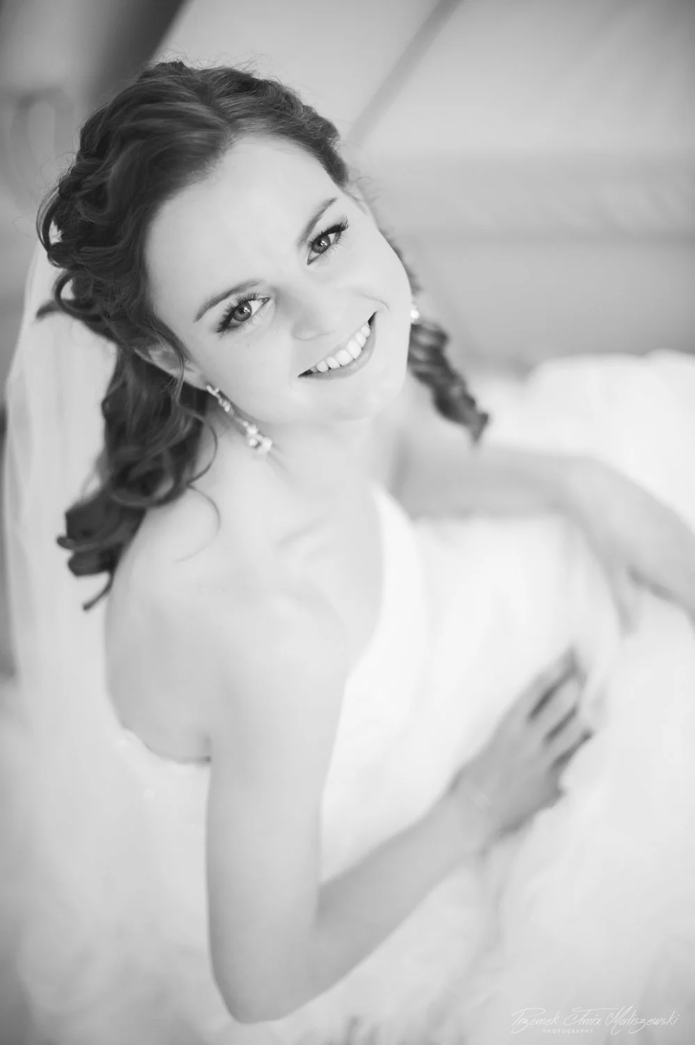Black and white photo of a smiling woman with curly hair, wearing earrings, in a strapless dress, looking up at the camera.