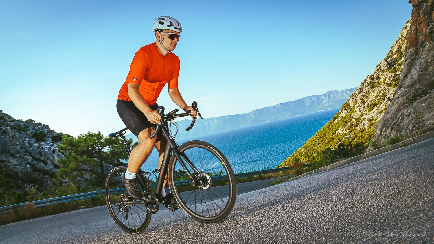A man in an orange cycling jersey and black shorts rides a black road bike along a coastal mountain road with blue ocean and distant mountains in the background.