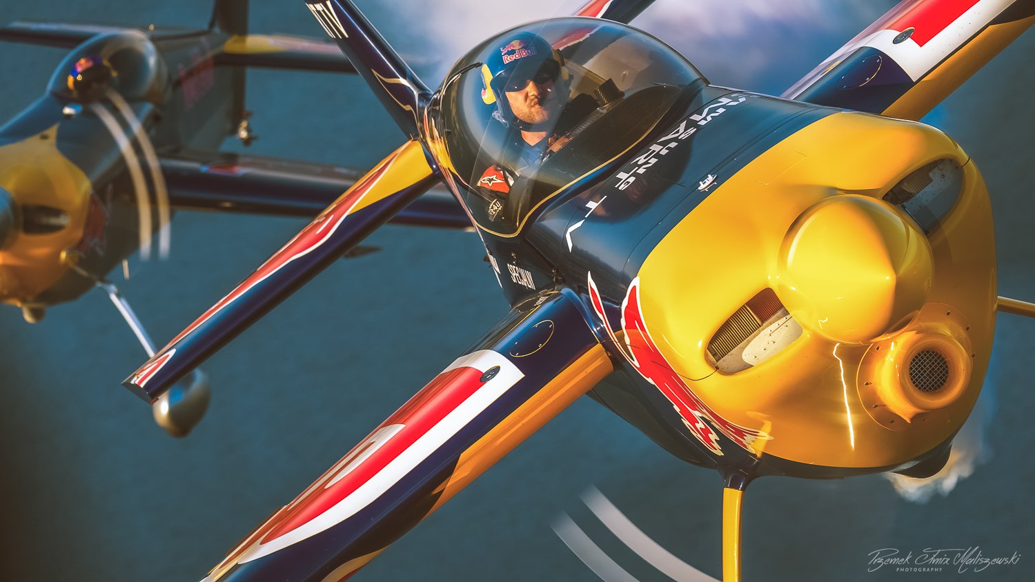 A fighter jet with a yellow nose and blue, red, and white body flying over water with a pilot wearing a helmet and sunglasses inside.