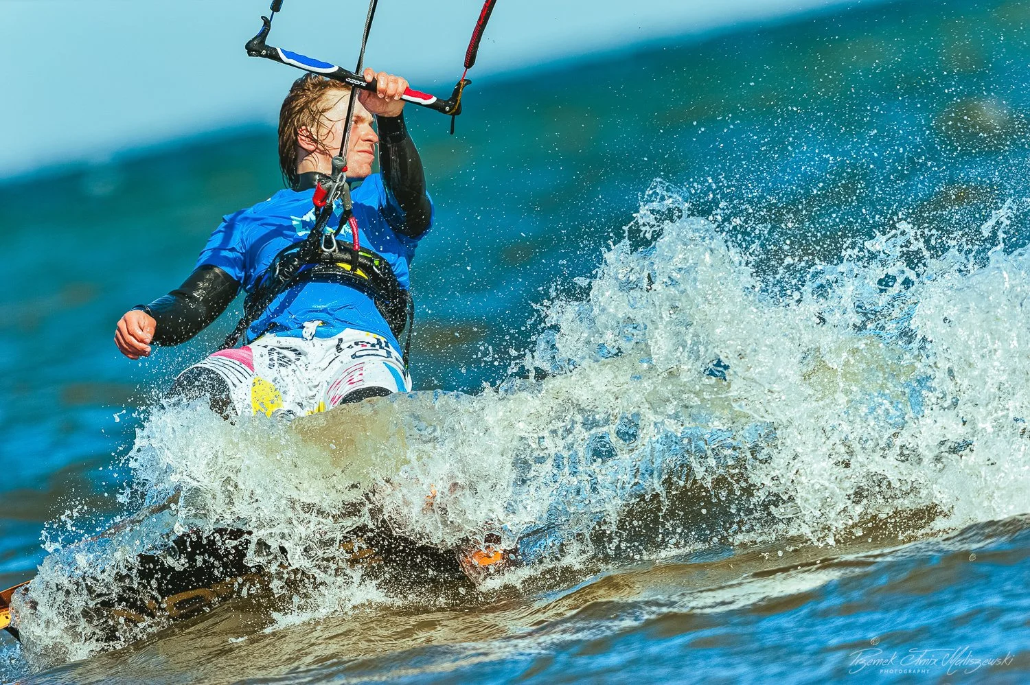 Person kiteboarding on the water, wearing a blue t-shirt and colorful shorts, holding onto the kite control bar with water splashing around.