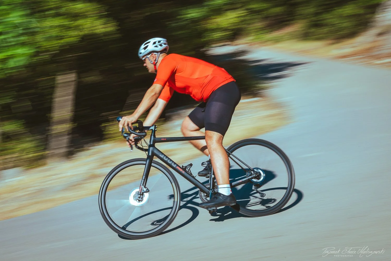 A man wearing a red shirt, black cycling shorts, and a helmet riding a black bicycle on a paved road through a wooded area.