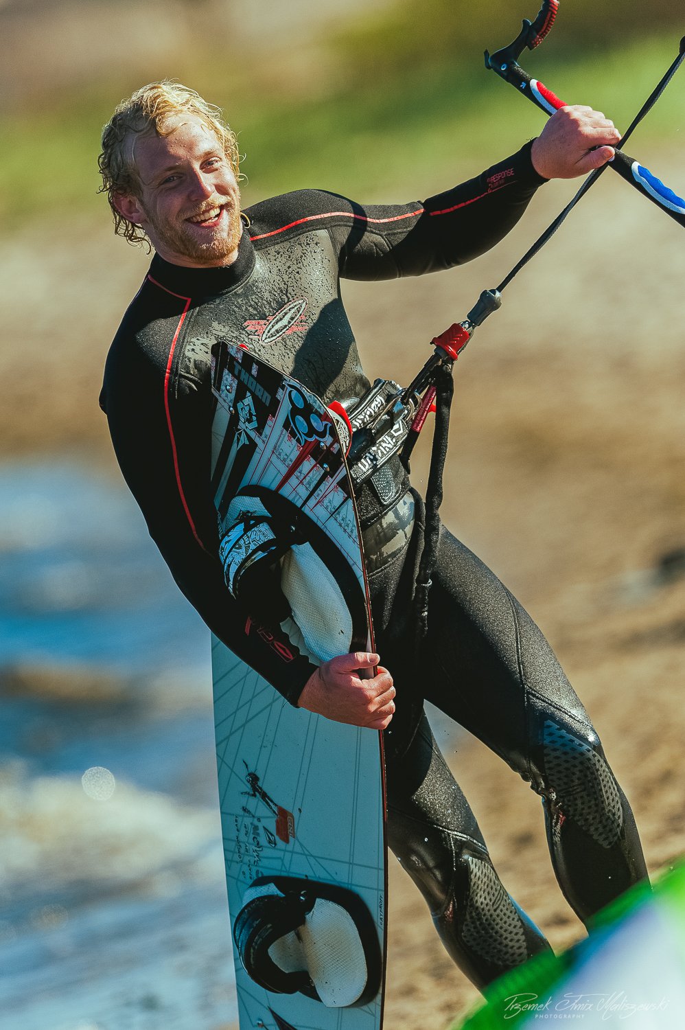 A man in a wet wetsuit holding a kiteboard and a kite control bar, smiling on a beach.