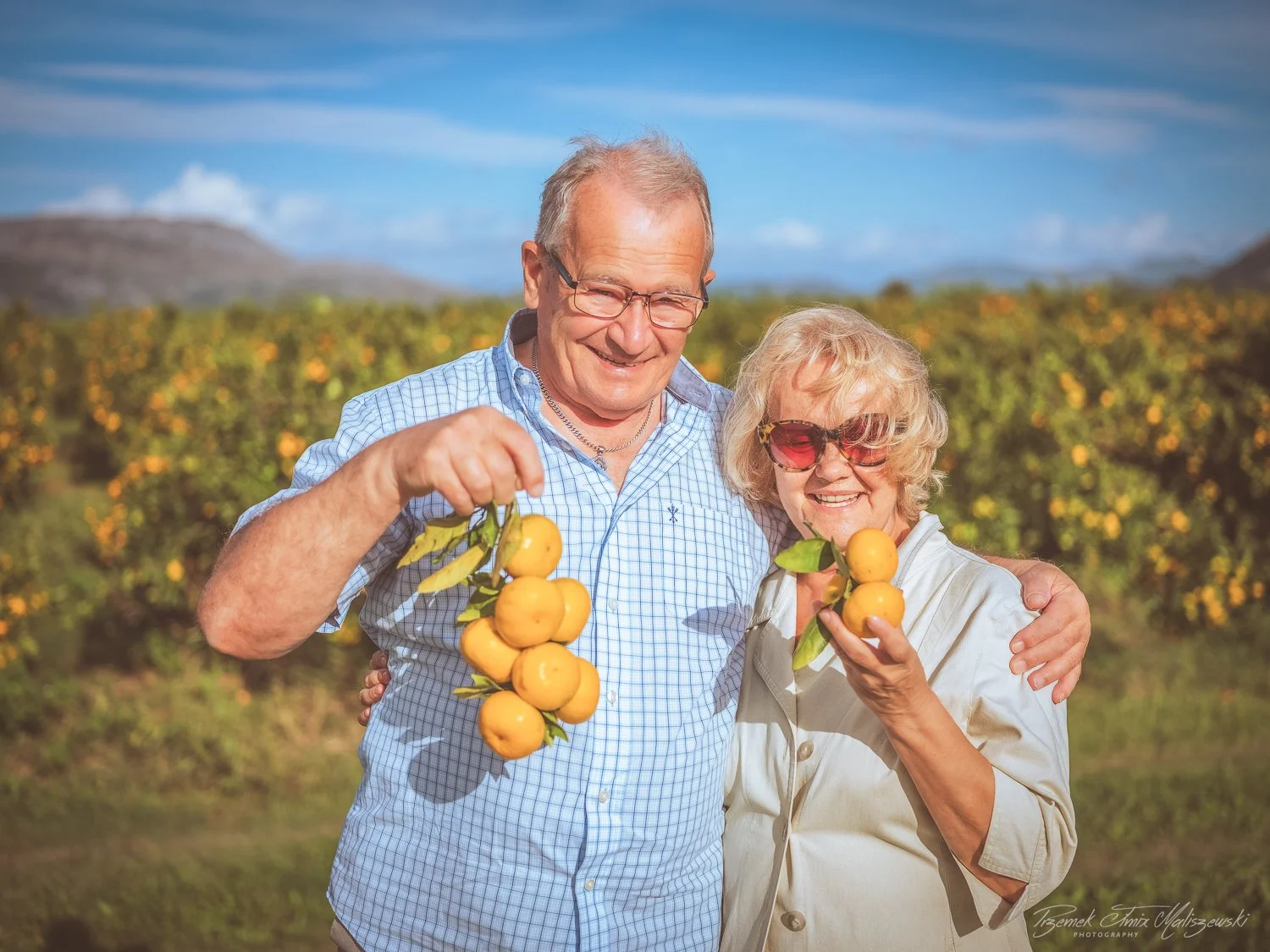 An elderly couple smiling and holding freshly picked kumquats in an orchard with a bright blue sky and distant mountains.