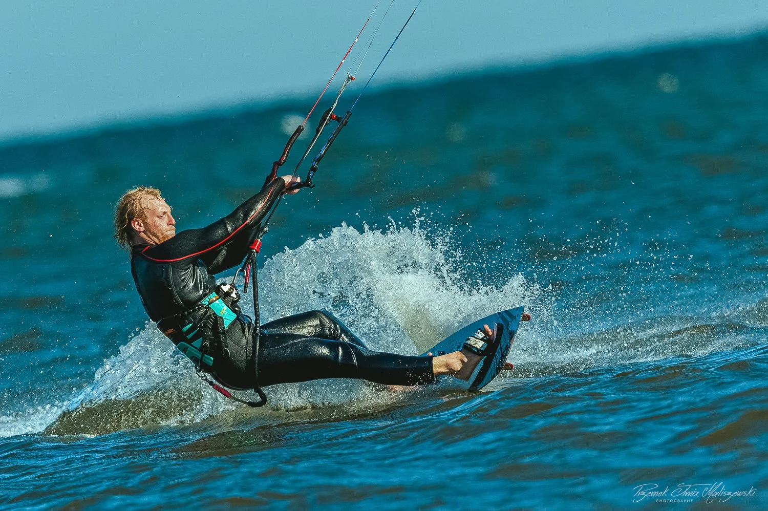 A man in a black wetsuit riding a kiteboard on the water, holding onto control bars connected to a kite, with water splashing around him.
