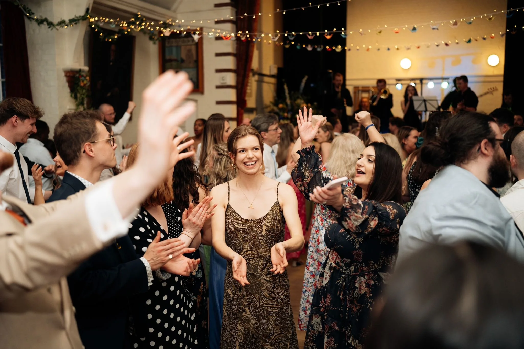 People dancing and celebrating at an event with a live band playing in the background, decorated with string lights.
