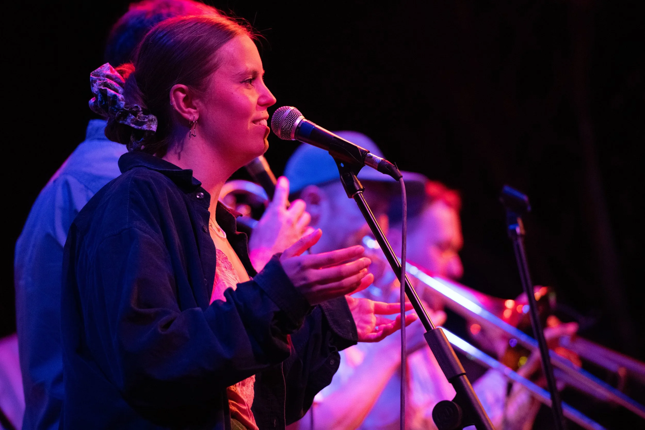 A woman singing into a microphone with musicians playing violins in the background, stage lighting with pink and purple hues.