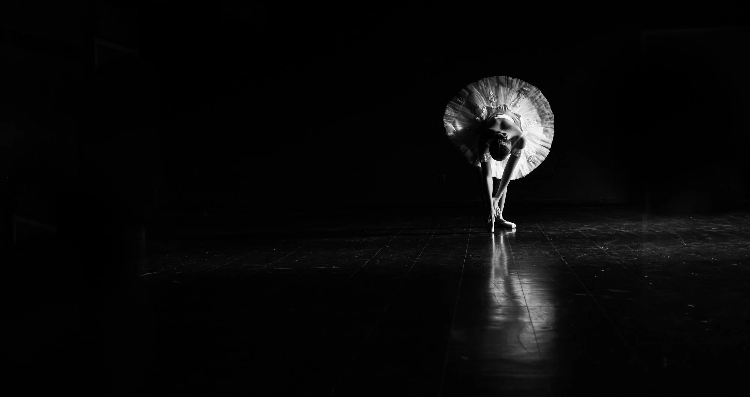 Ballet dancer in a tutu performing on stage, bending forward with arms reaching toward the floor, illuminated by a spotlight in a dark theater.