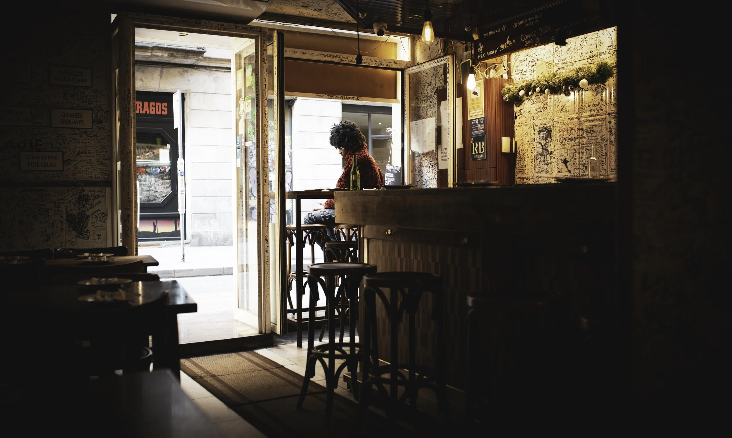 A person with curly hair sitting at a bar table inside a cozy cafe, looking out onto the street through an open door. The cafe has wooden furniture, decorated walls with graffiti and a garland, and warm lighting.