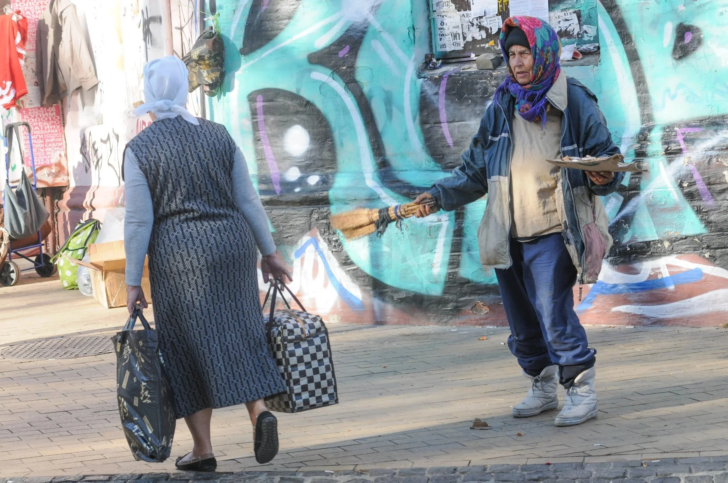 A woman in a long dress and headscarf carries shopping bags and a suitcase as she walks past a man in casual clothing who is offering her a broom and holding a plate, standing in front of a graffiti-covered wall.
