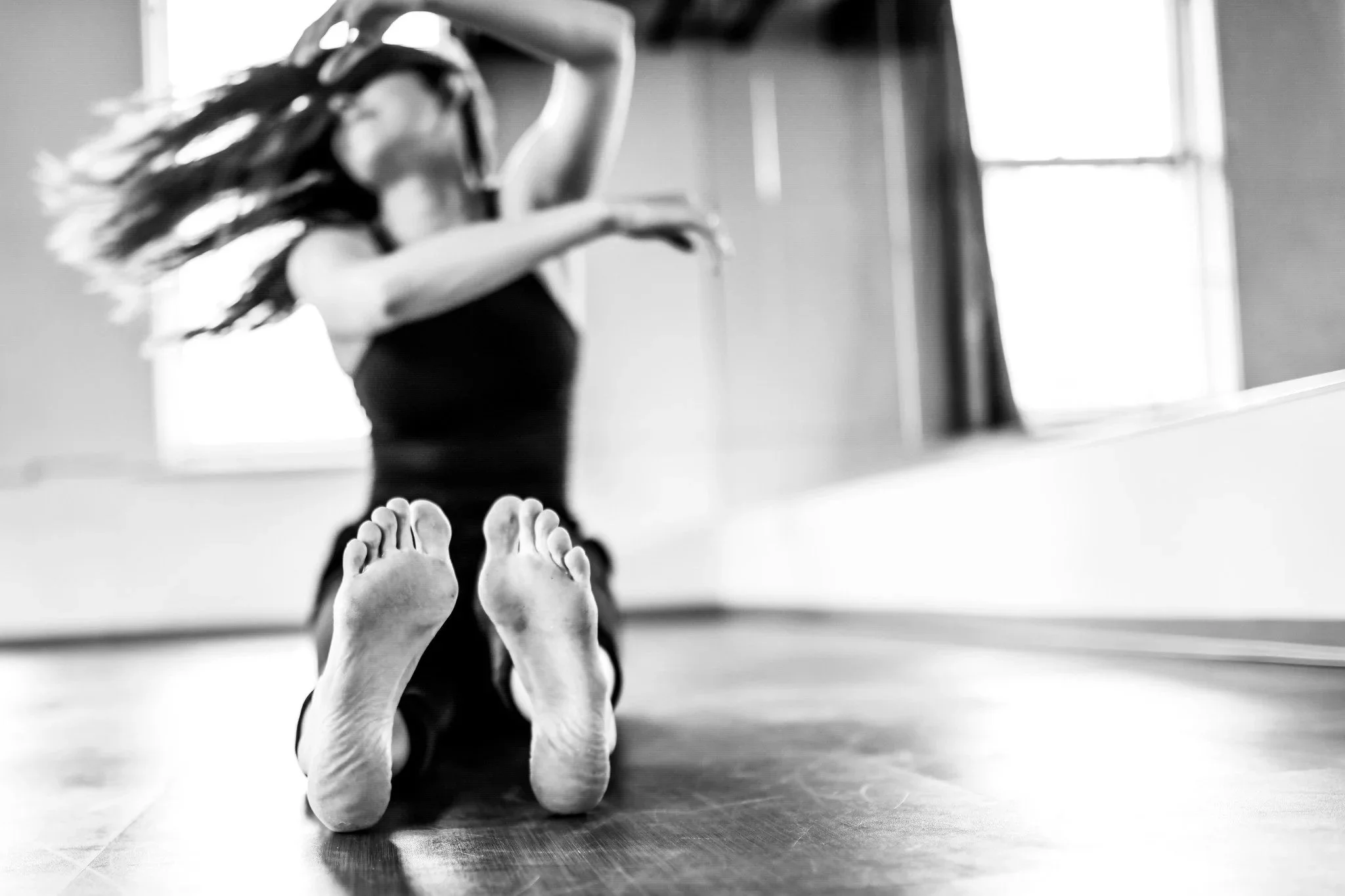 A person with long hair is doing a dance pose on a wooden floor in a studio, with a large window letting in natural light.