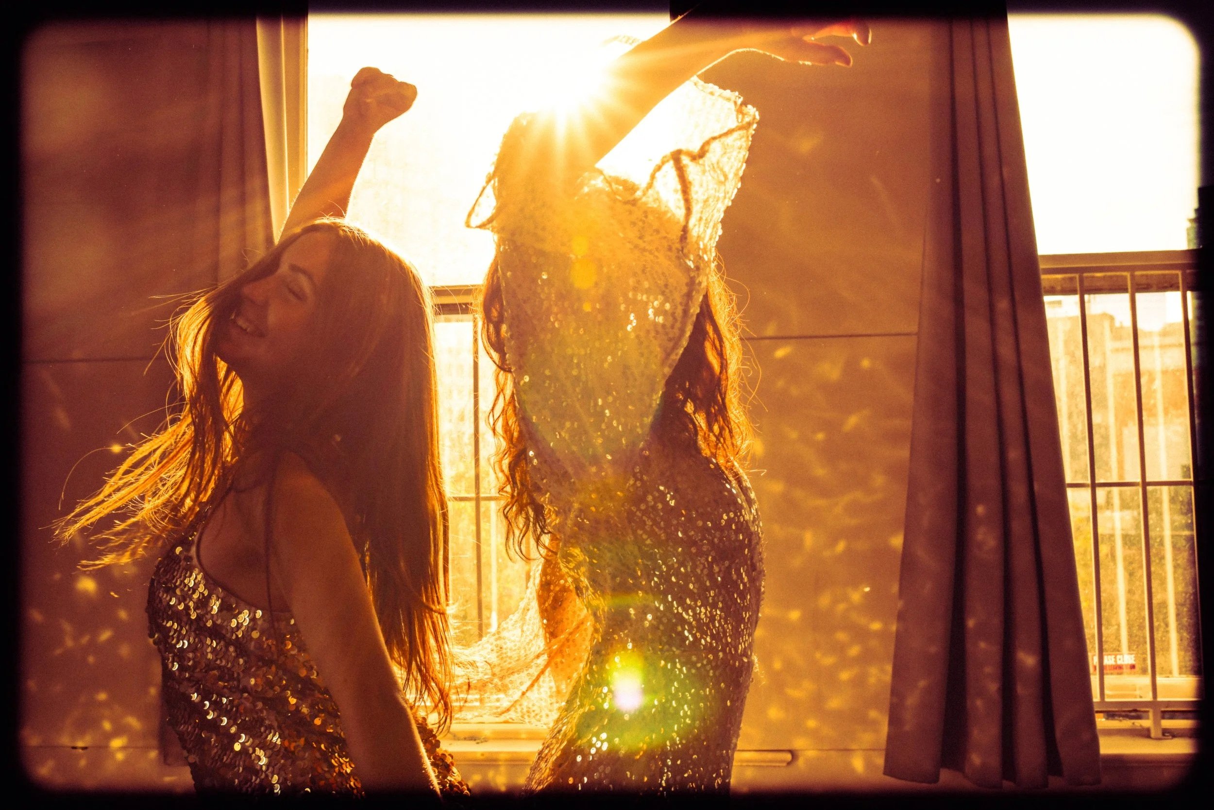 Two women dancing in front of a window with sunlight shining through, wearing shiny, sequin dresses.