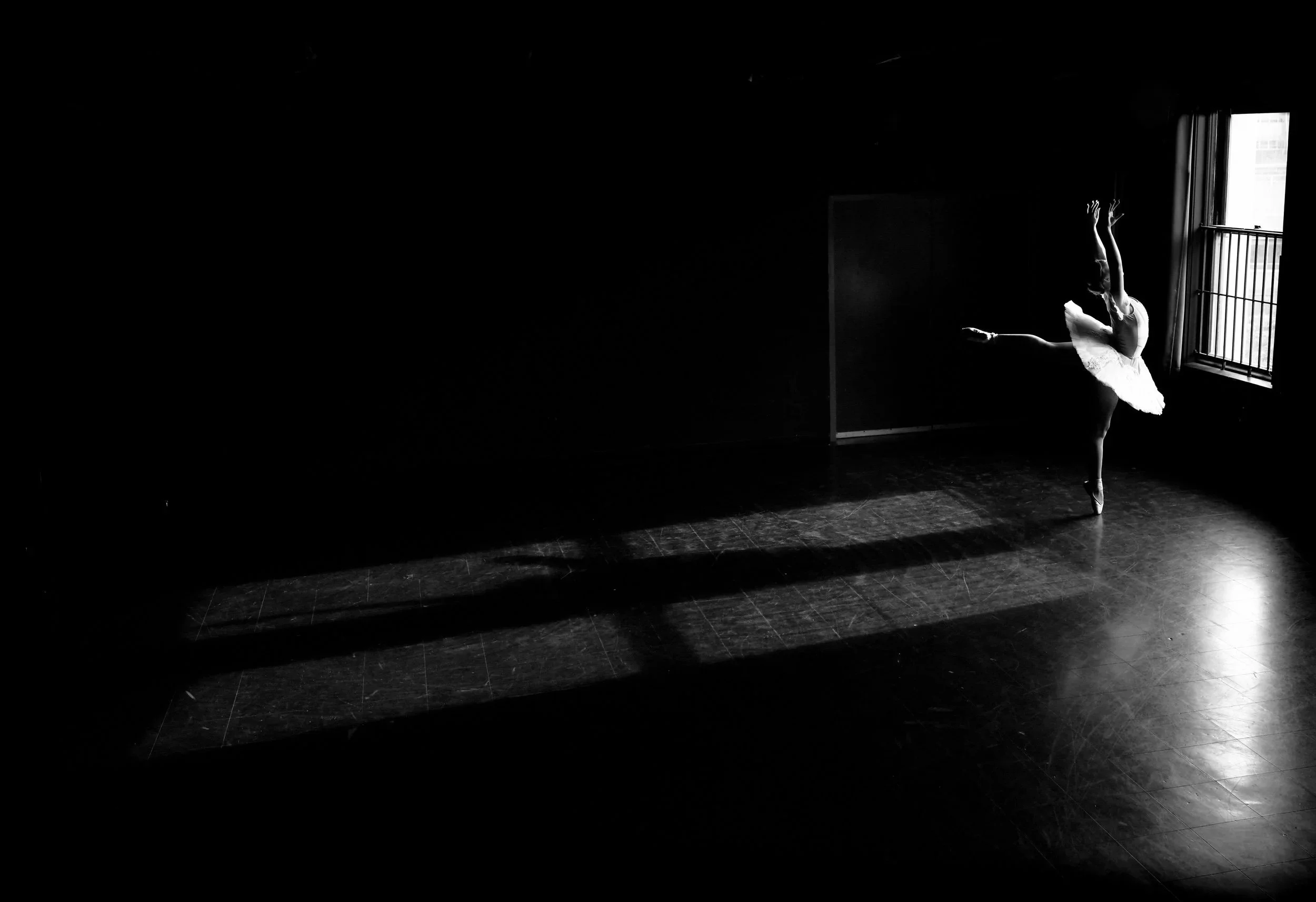 A ballet dancer practicing alone in a dimly lit studio near a window, wearing a tutu and balancing on one leg with arms raised.