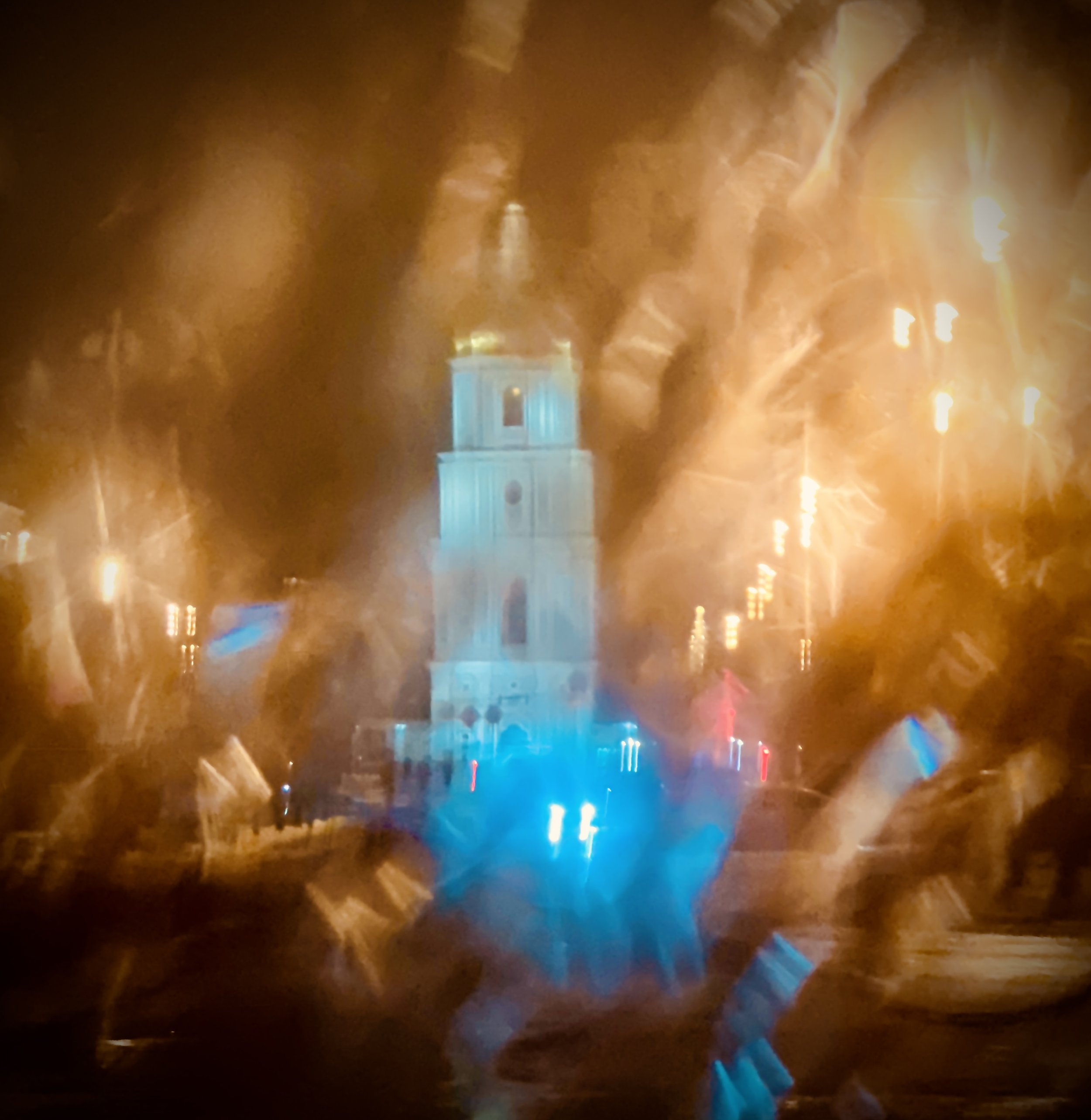 Nighttime view of a church tower illuminated in blue, seen through a rainy window with streaks and droplets creating a blurred, distorted effect.
