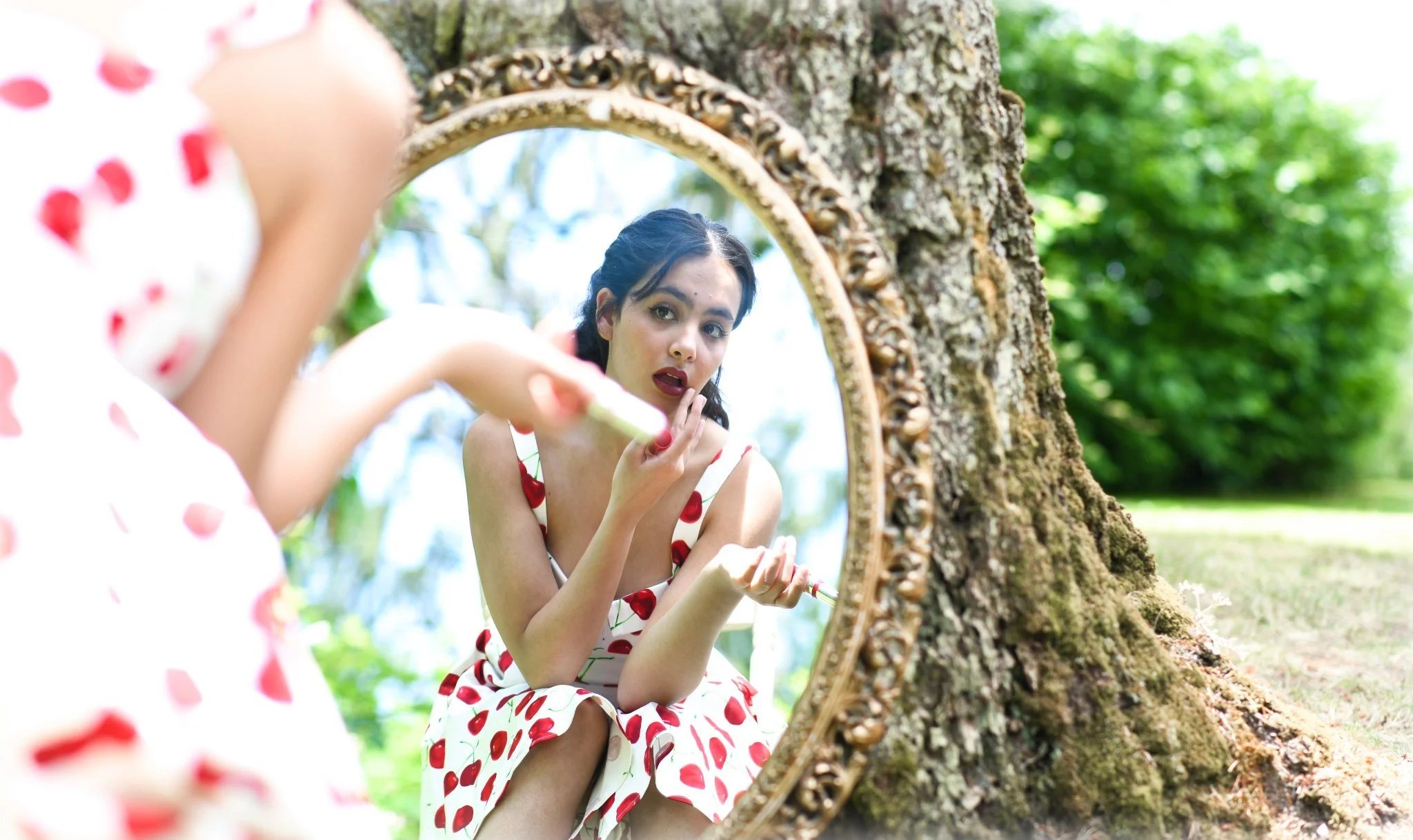 Woman in a dress with cherry patterns applying makeup while looking into a mirror outside, with the mirror propped against a tree trunk.