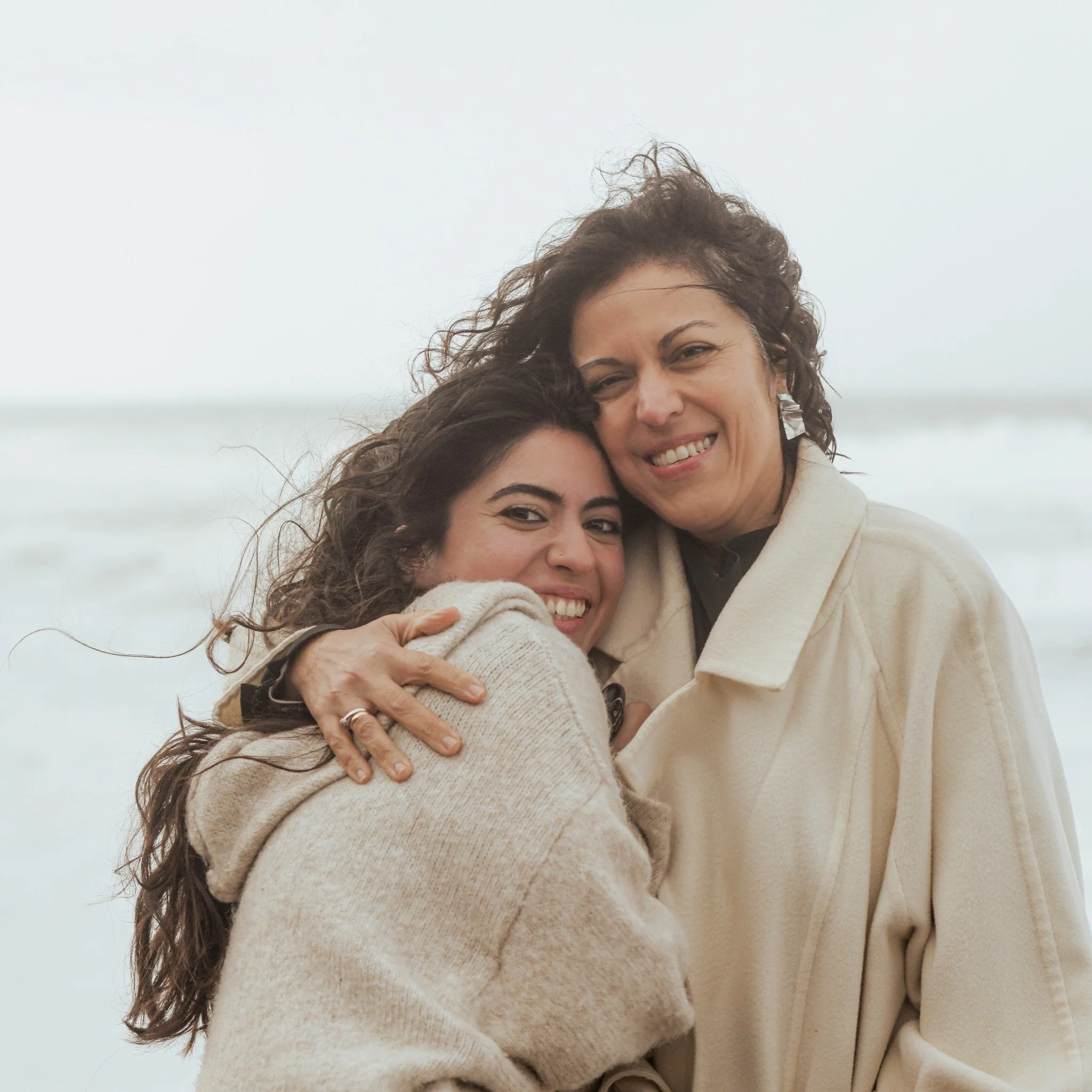 Two women hug and smile at the camera on a beach with overcast sky in the background.