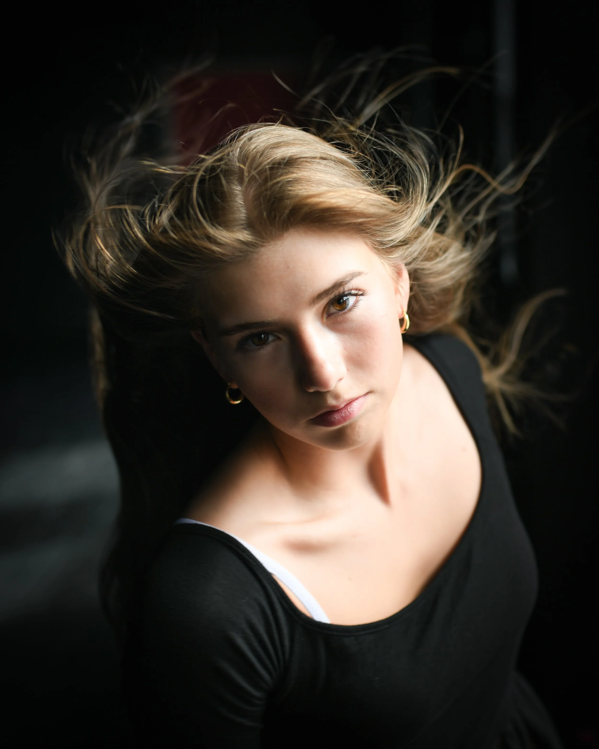 Portrait of a young woman with flowing blonde hair, wearing a black top and gold earrings, looking directly at the camera against a dark background.