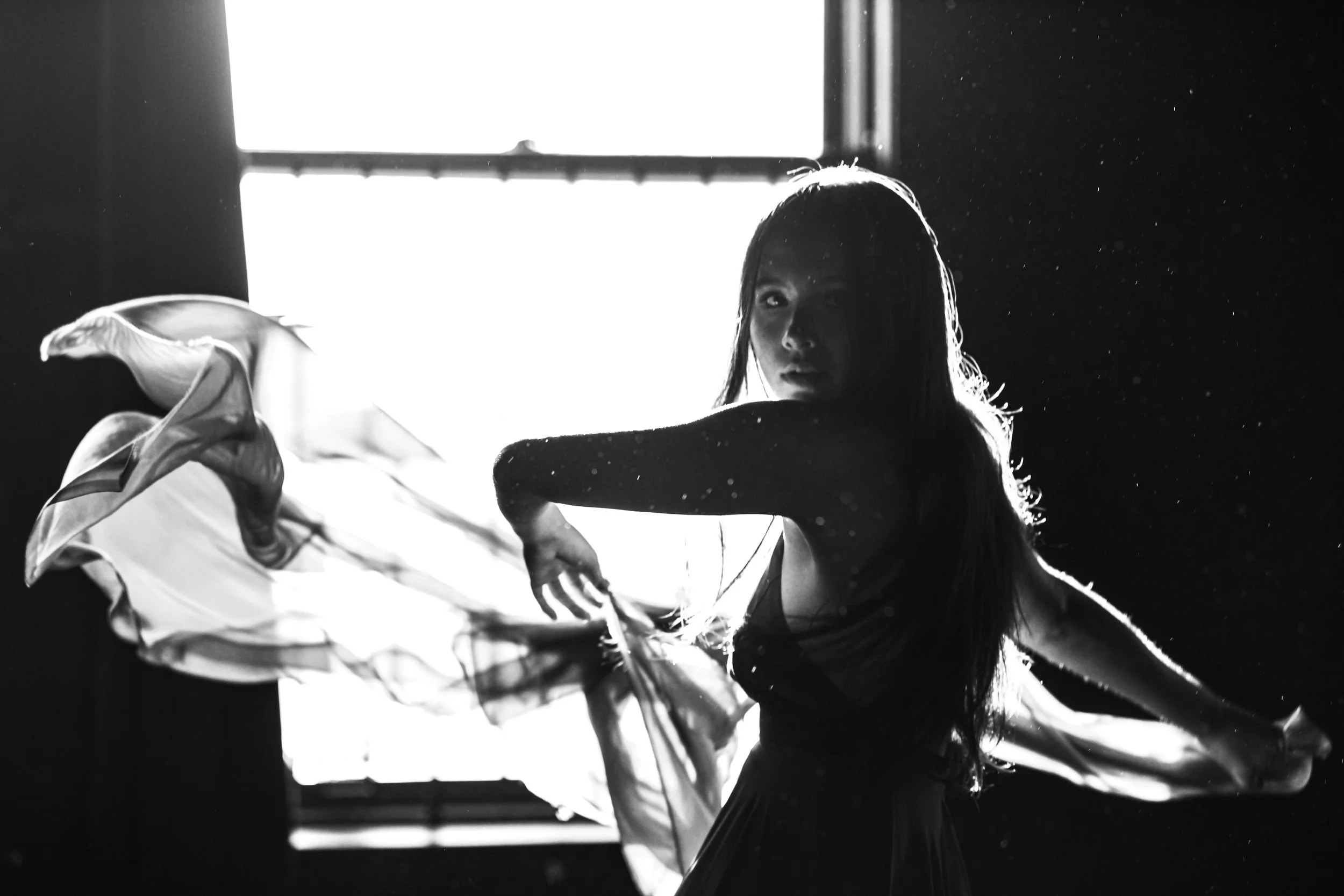 A woman with long hair poses in front of a bright window, creating a silhouette effect with light and shadow, holding flowing fabric.