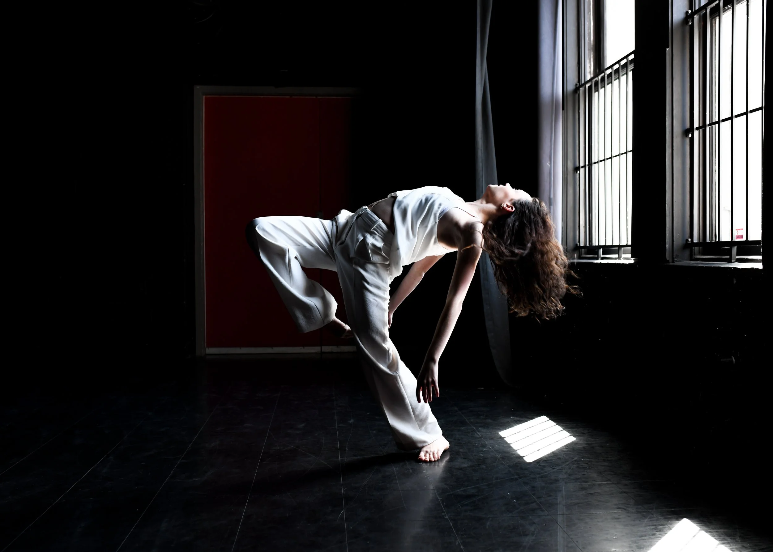 A woman practicing yoga in a dimly lit studio near large windows, balancing on one leg and stretching her upper body backward.
