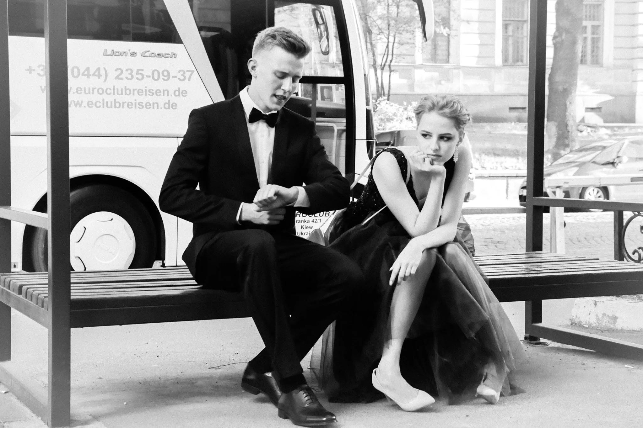 A man in a tuxedo and a woman in a fancy dress sitting on a park bench, with a tour bus in the background.