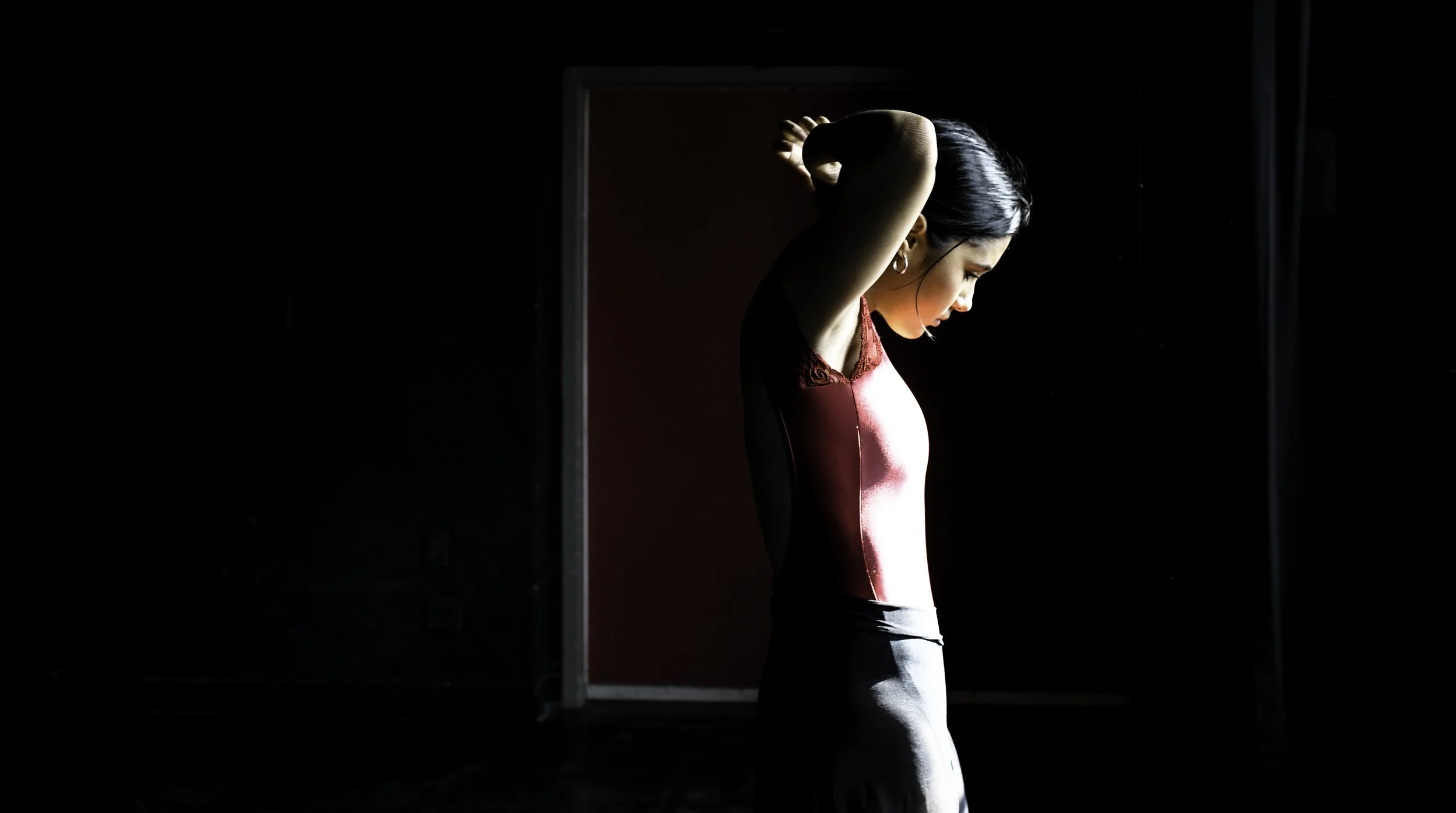 A woman in a red and black dress stands in profile with her arms raised behind her head in a dark room, illuminated by light from a doorway.